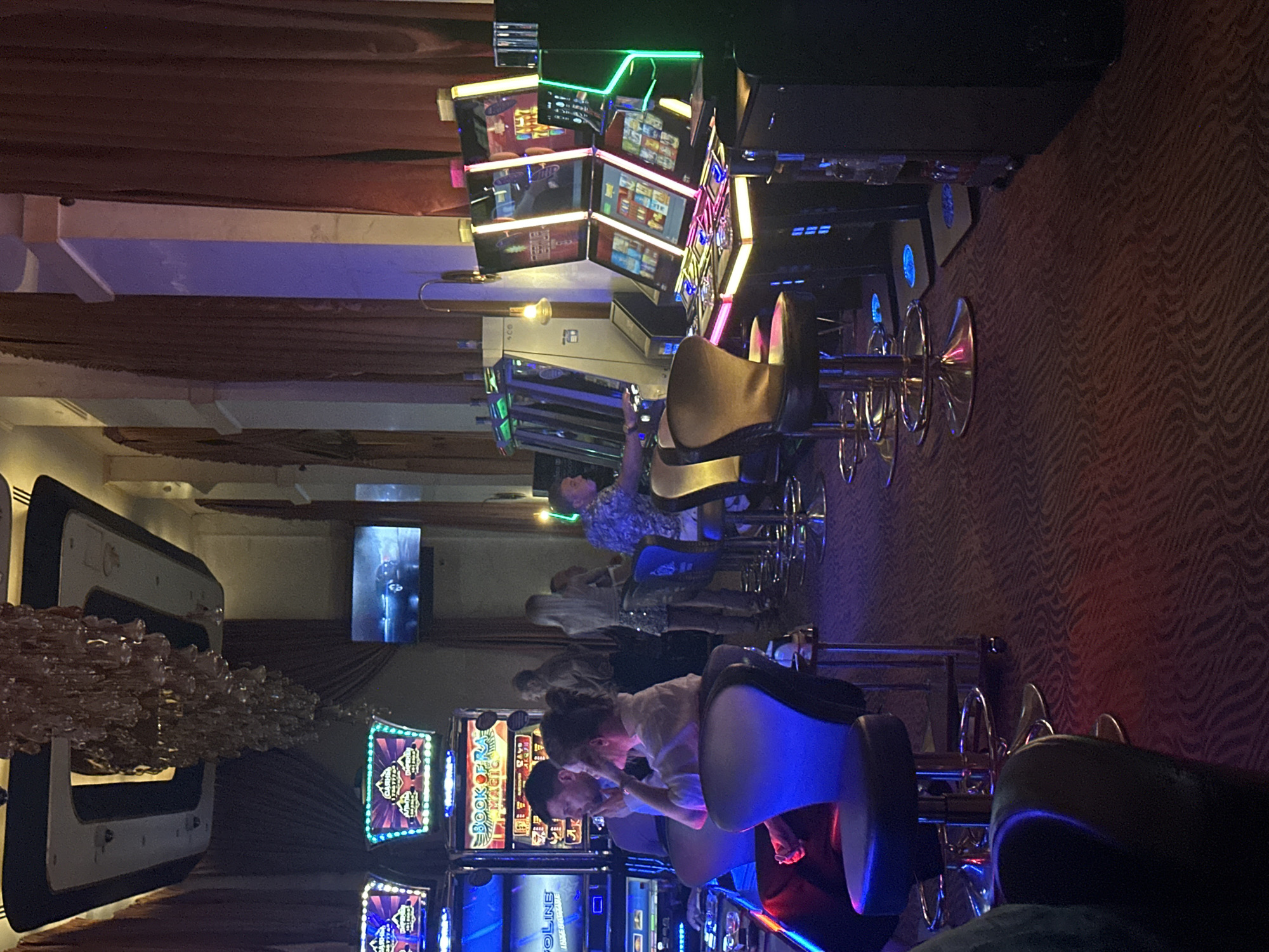 The image captures an interior scene within a casino, likely located in Estosadok, Russia, based on its context. The environment is dimly lit, with prominent neon and LED lighting emanating from rows of slot machines. The casino floor is carpeted with a dark, patterned rug.
In the foreground, several individuals are seated at or near slot machines. To the left, a woman with dark hair pulled back is seated in a blue upholstered stool, facing away from the camera and towards a slot machine. Beside her, a man with dark hair is seated in a similar stool, also facing a slot machine and appearing to be engaged with it. Further to the right, other patrons are visible, some seated at machines and others standing. One woman with blonde hair is standing and looking towards the slot machines. A man in a patterned shirt is seated at a slot machine on the right side of the image.
The slot machines themselves are modern, with illuminated screens displaying game graphics and information. The machines vary in design and color, with some featuring prominent neon accents. In the background, a large, elaborate chandelier hangs from the ceiling, contributing to the opulent atmosphere of the casino. A flat-screen television is mounted on the wall in the mid-ground, displaying an image of a car. Heavy curtains, likely red or burgundy, line the walls, adding to the enclosed and somewhat theatrical feel of the space. The overall impression is one of a bustling, brightly lit casino environment.