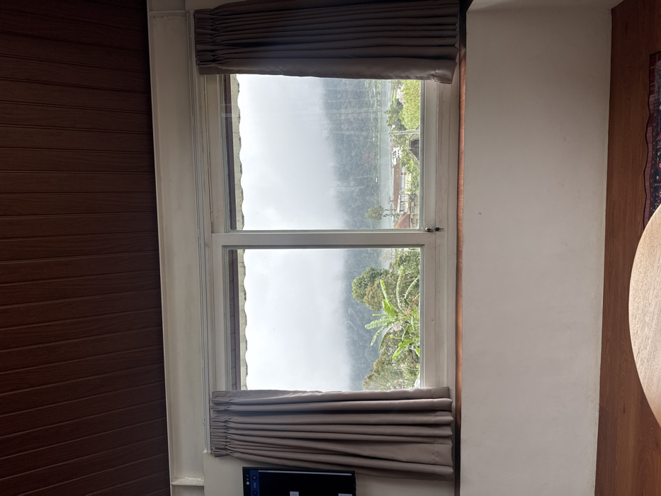 A double-paned window reveals a misty, mountainous landscape. The upper pane shows a foggy view with indistinct buildings. The lower pane features green foliage, including banana trees. Gray curtains frame the top and bottom of the window. The window is set between a wall with brown vertical paneling on the left and a plain white wall on the right. A dark electronic device is partially visible at the bottom center, below the lower curtain.