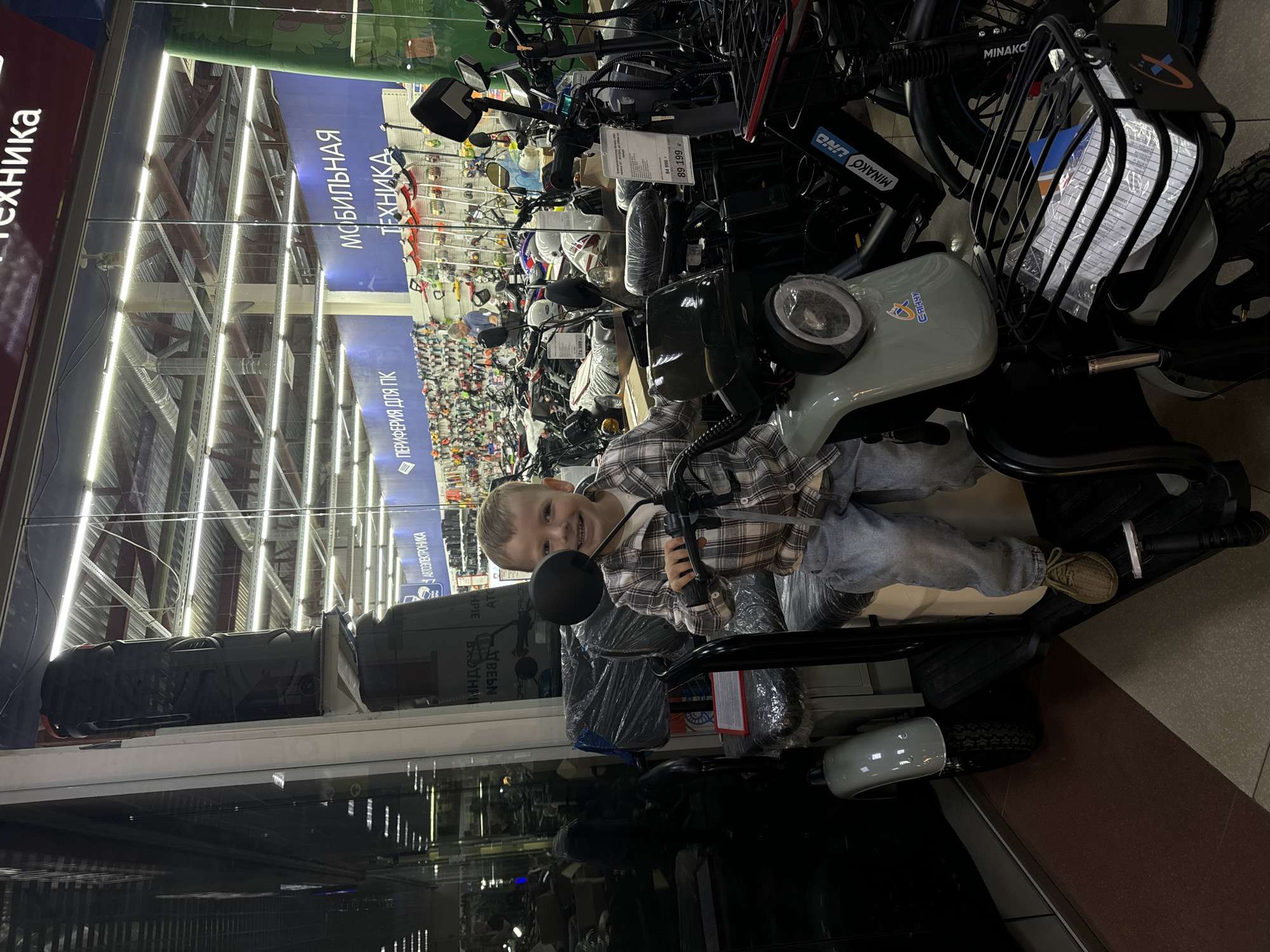 A young boy is sitting on a light gray electric scooter in a store. He is wearing a plaid shirt and blue jeans. The scooter has a black seat, a black handlebar with mirrors, and a front basket containing papers. In the background, there are more scooters and merchandise, with signs indicating "Мобильная техника" (Mobile Technology) and "Периферия для ПК" (PC Peripherals). The store appears to be well-lit with fluorescent lights. The floor has a tiled pattern. The setting is the "Технодом" store located in the city of Sharya, Russia.