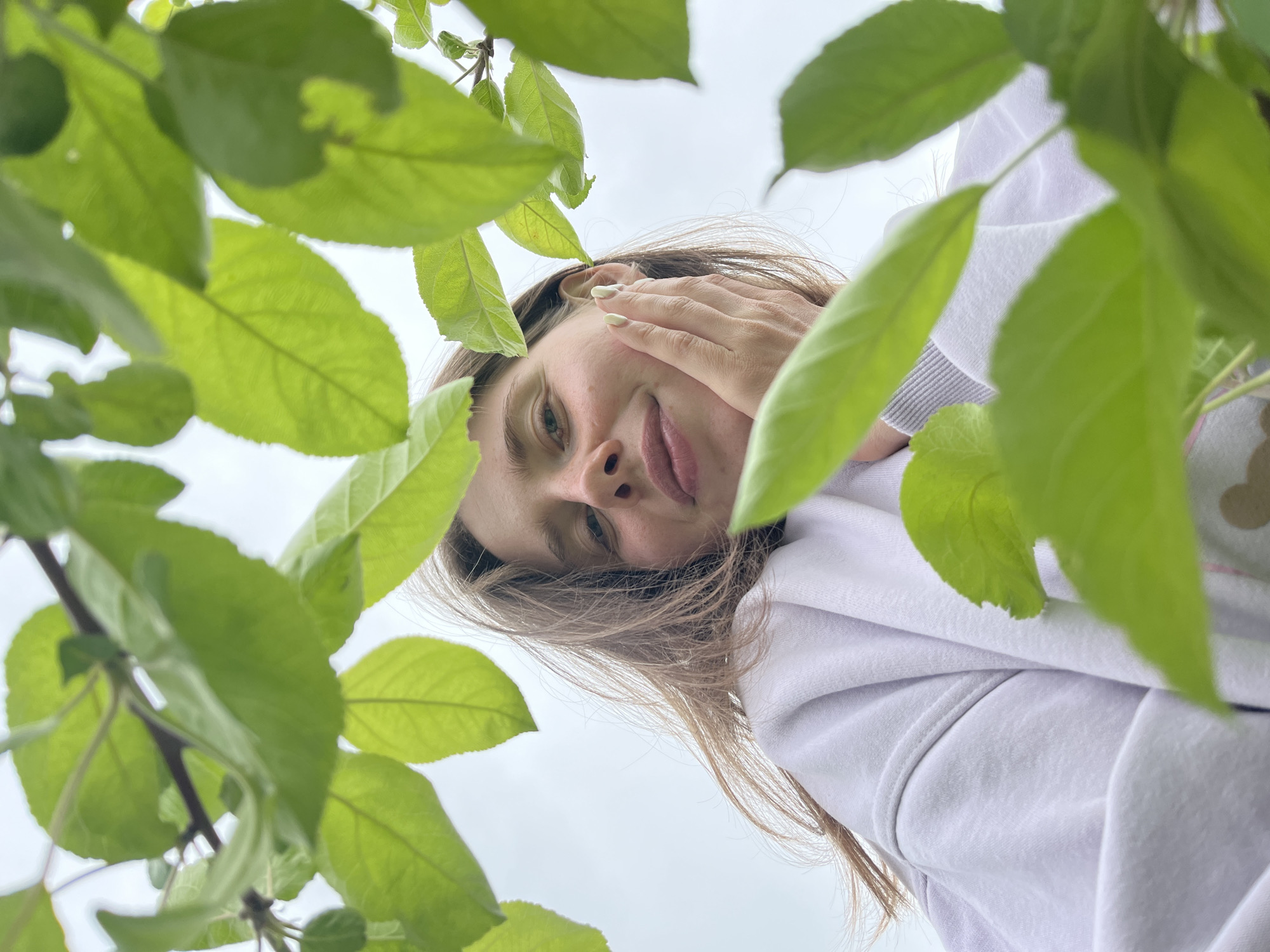 The image depicts a young woman looking down at the camera from beneath a tree. She is fair-skinned with brown hair and is wearing a white hoodie. Her right hand is raised to her face, partially obscuring her cheek. The background is the sky, partially obscured by the green leaves of the tree. The location is outdoors, though specific details about Chaikovsky, Russia are not evident in the image.