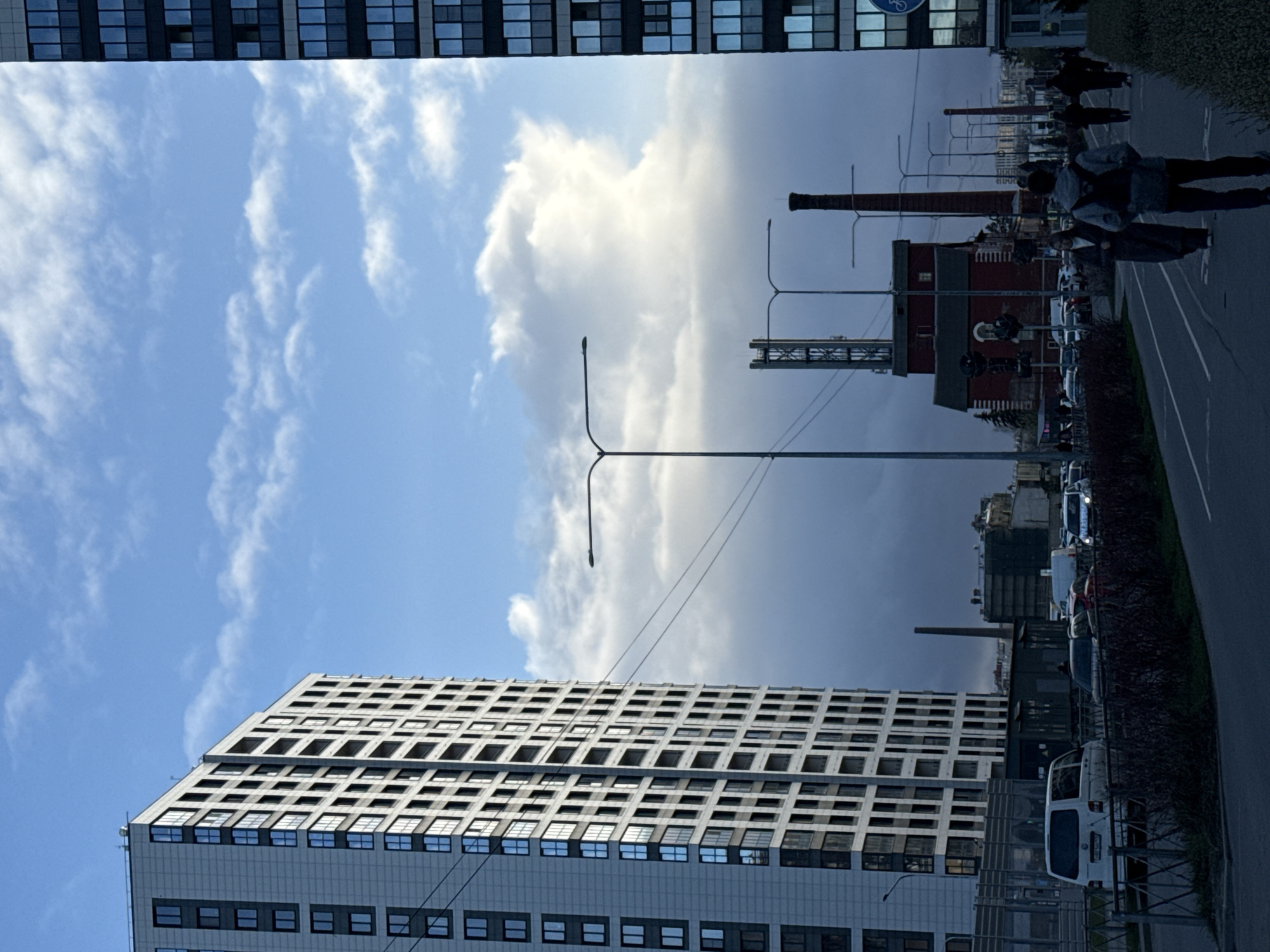 The image shows a street scene in Saint Petersburg, Russia. A tall, modern apartment building with a grid-like facade of windows is on the lower left. Overhead power lines run through the scene. In the background, a red brick industrial building with a tall chimney is visible. A street with parked cars and a sidewalk is on the right. People are walking on the sidewalk. The sky is partly cloudy, with a mix of bright blue sky and dark rain clouds.