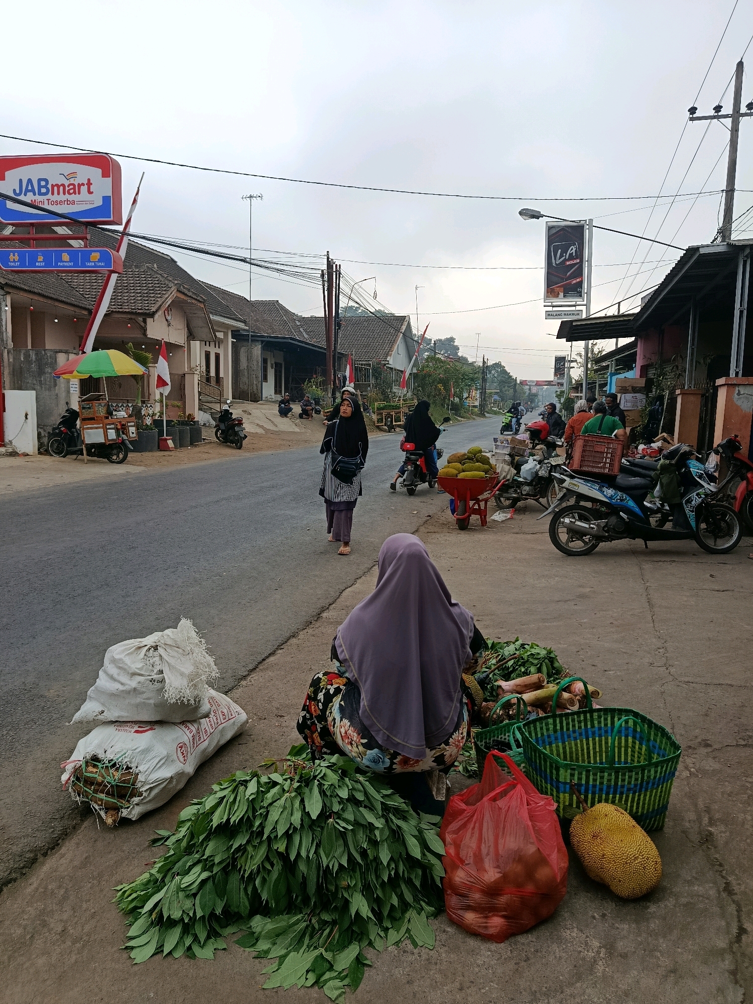 The image depicts a street-side market scene in Tajinan, Indonesia, under an overcast sky. In the foreground, a person wearing a purple headscarf and a floral patterned skirt is seated on the ground, surrounded by produce. Several bunches of green leafy vegetables are spread out before them, along with two large white sacks, one of which appears to contain bundled wood. Adjacent to the vegetables are two green woven baskets, a red plastic bag filled with what appear to be fruits, and a ripe jackfruit.
Further back, a street runs parallel to the market stalls. Several motorcycles are parked along the side of the road. Two women wearing hijabs are walking on the sidewalk. One woman, in the middle ground, is wearing a black hijab and a striped shirt, carrying a dark bag over her shoulder. The other woman is slightly behind her.
On the left side of the street, a store sign for "JABmart Mini Tosserba" is visible. Next to it, a small food cart with a colorful umbrella is set up, with a motorcycle parked nearby. On the right side of the street, a tall sign displaying "LA" with text below it stands next to a building. Several other people are visible in the background, some sitting on the ground and others near motorcycles. The environment appears to be a typical street in a town or city, with buildings lining the road and utility poles with electrical wires overhead. The overall atmosphere suggests an early morning or late afternoon market activity.