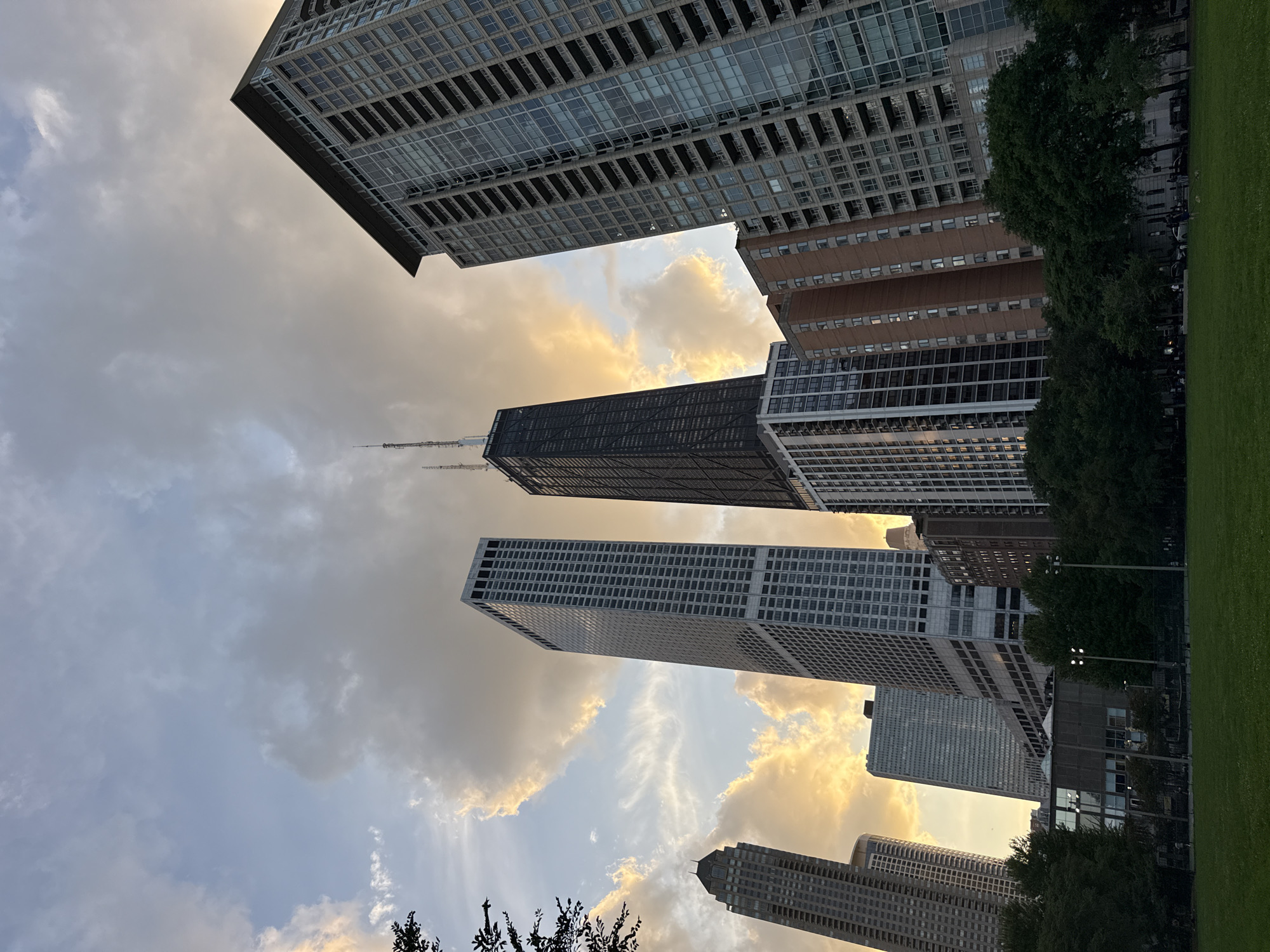 The image captures a view of the Chicago skyline with a focus on several prominent skyscrapers. The John Hancock Center, characterized by its dark, crisscrossed exterior, is a central feature. To its left is a tall, slender, white skyscraper with a grid-like facade, likely 875 North Michigan Avenue. To the right, a large, modern residential building with balconies dominates the frame. In the foreground, a lush green lawn stretches out, hinting at a park setting. Trees line the mid-ground, and further back, tennis courts and some people can be observed. The sky above is filled with dramatic clouds illuminated by the warm tones of a sunset or sunrise, creating a striking contrast with the urban architecture. The overall impression is one of urban grandeur set against a beautiful natural sky.