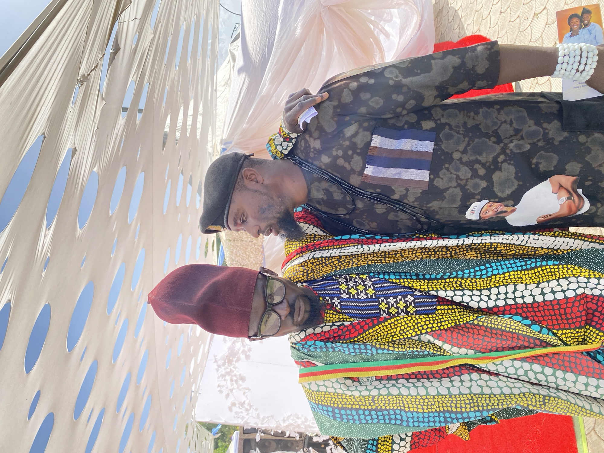 Two men are standing close together, with one partially overlapping the other. The man on the left wears a burgundy cap and glasses. He is draped in a colorful shawl-like garment with patterns in various colors including red, yellow, green, and blue. The man on the right wears a black beret and has facial hair. His outfit is dark with lighter, mottled patterns. He is also wearing white beaded bracelets and a black beaded necklace. The background is a mix of a white canopy-like structure with numerous holes, and glimpses of the sky. A small picture of a smiling couple and a cut out picture of a man wearing a white hat and watch are pinned on the right man's garment. The location is Jos, Nigeria.