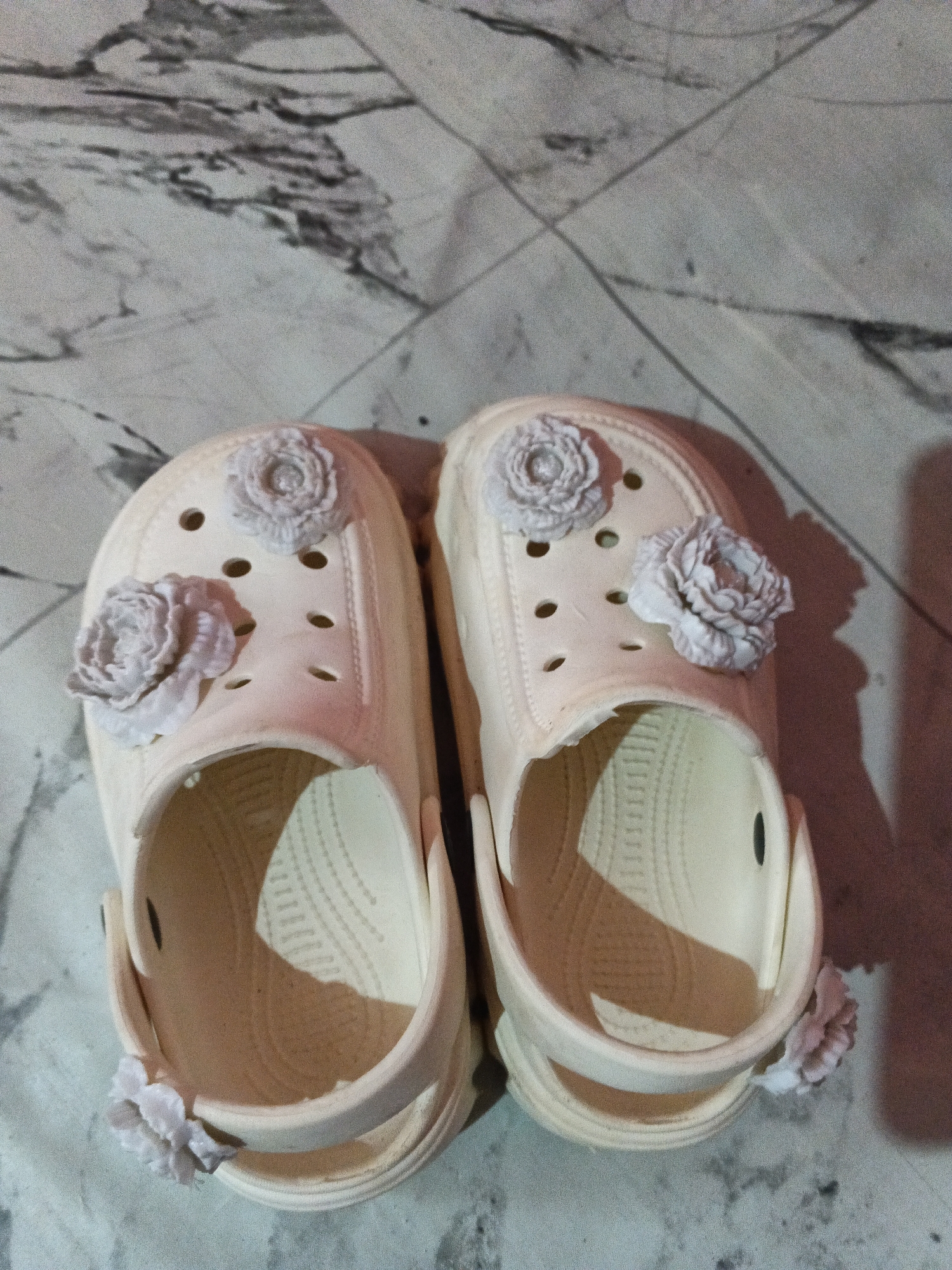 A pair of light beige clogs adorned with light gray flower embellishments rests on a gray and white marbled floor. The clogs are positioned side-by-side, facing upwards, with the open interiors visible. Each clog features two flower charms on the top surface, and one on the back strap. The scene is illuminated by artificial light, casting shadows on the floor. The background consists of a marbled surface, suggesting an indoor setting. There are no people or visible actions occurring in the image.