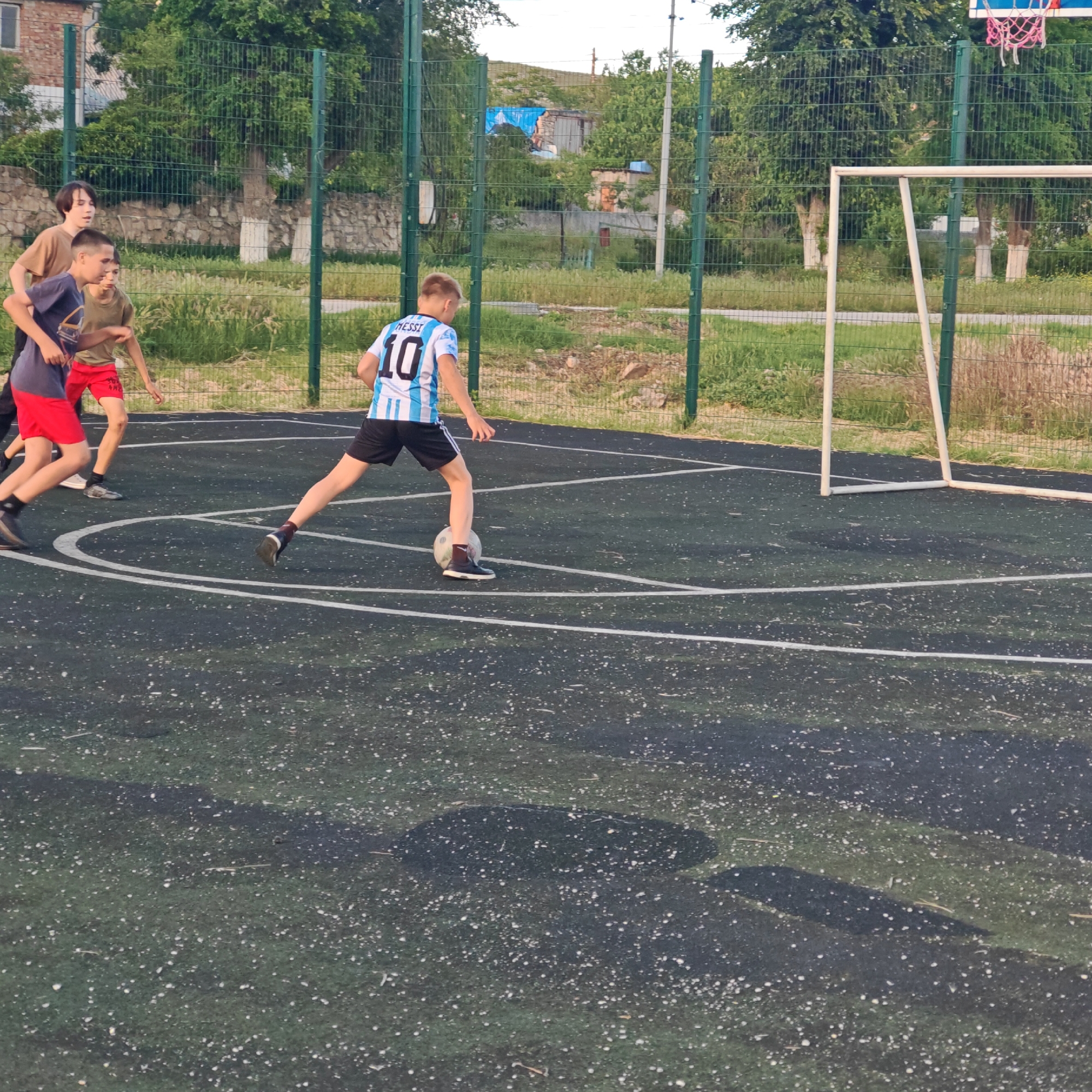 On a sports court in Мысовское сельское поселение, Ukraine, four young males are engaged in a soccer game near a white-framed goal. One boy, wearing a light blue and white jersey with "Messi 10" printed on the back, is actively dribbling the soccer ball. The other three boys are nearby, presumably participating in the game as well. A green fence surrounds the court, separating it from the surrounding area. A basketball hoop is visible in the background. The court surface is dark green with white lines marking out the boundaries of the playing area. Trees and some buildings are visible in the background.