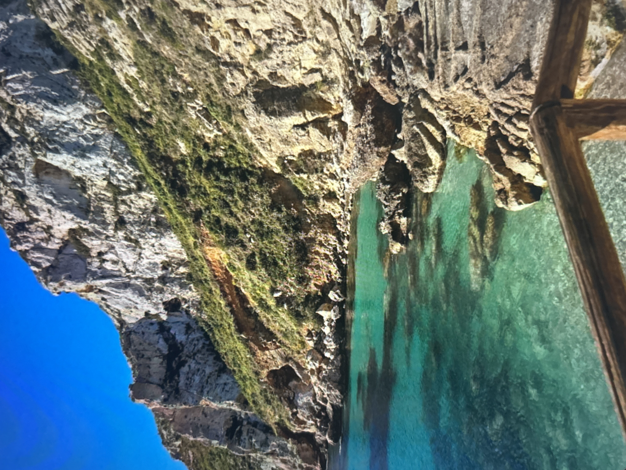 The image displays a vibrant coastal landscape featuring majestic cliffs and incredibly clear turquoise water, oriented vertically but depicting a horizontal scene. The left side of the frame is dominated by towering, rugged rock formations, varying in color from light grey to earthy browns and yellows. Patches of lush green vegetation cling to the cliff faces, indicating life on the steep terrain.
The right side of the image features a serene body of water with remarkable clarity, revealing the rocky seabed below. The water transitions from a bright aquamarine near the shore to deeper blues and greens further out. A wooden structure, possibly a railing or part of a pier, is visible in the far right foreground, suggesting a viewpoint or access point to the water.
The sky, a brilliant, cloudless blue, is visible in the upper left corner, indicating a clear, sunny day. No people or animals are present, emphasizing the natural beauty and tranquility of the location. The scene evokes a sense of pristine wilderness and natural grandeur, typical of a secluded cove or bay. There is no visible text in the image.