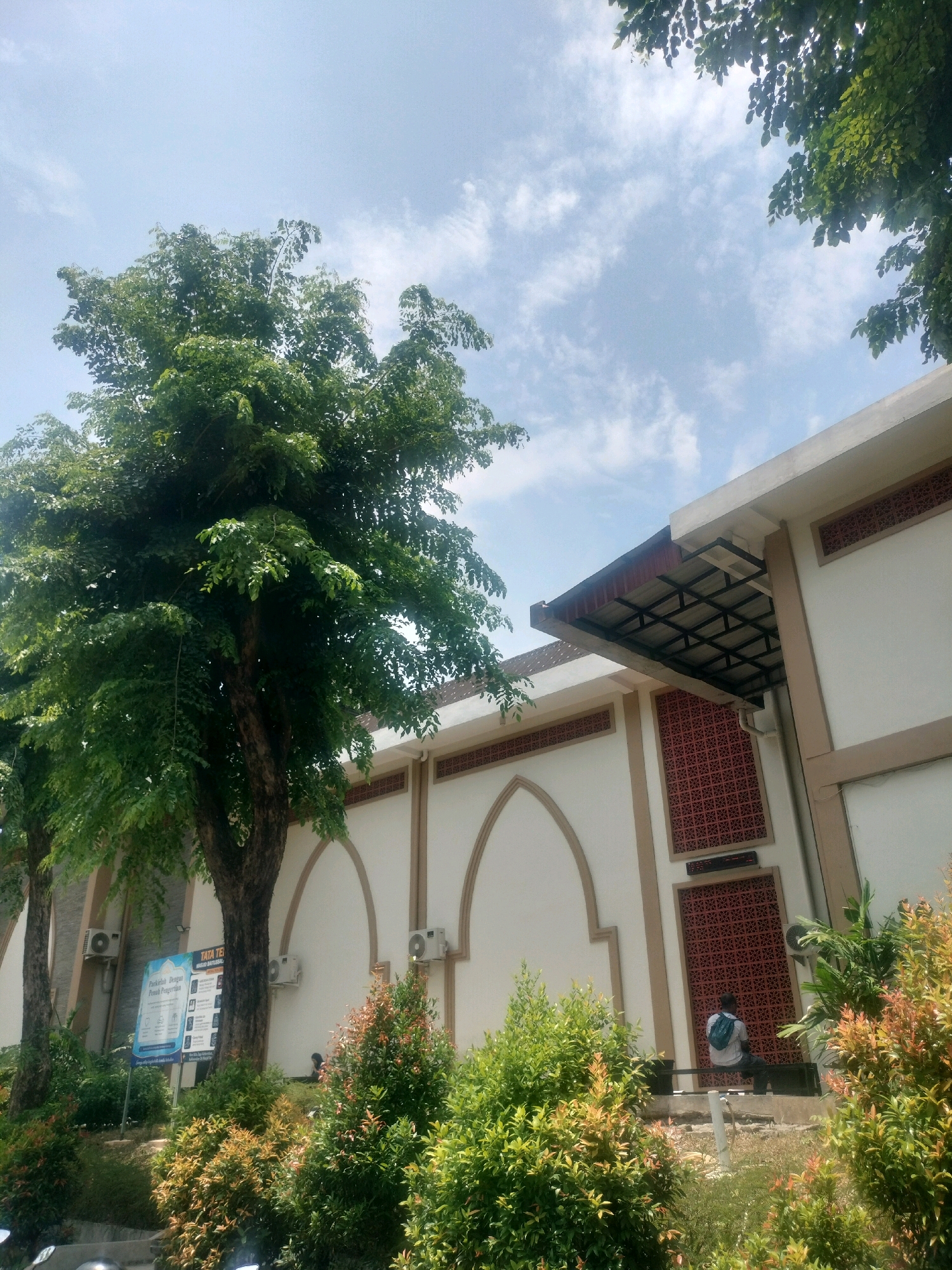 The image captures a view of the entrance area of the Batam City mosque. A person is seen sitting on a bench outside the mosque, wearing a light-colored shirt and a backpack. The mosque's exterior features a light-colored facade with decorative patterns and arched windows. Lush green trees and bushes surround the mosque, with the sky visible above. The scene is bathed in daylight, suggesting it was taken during the day.