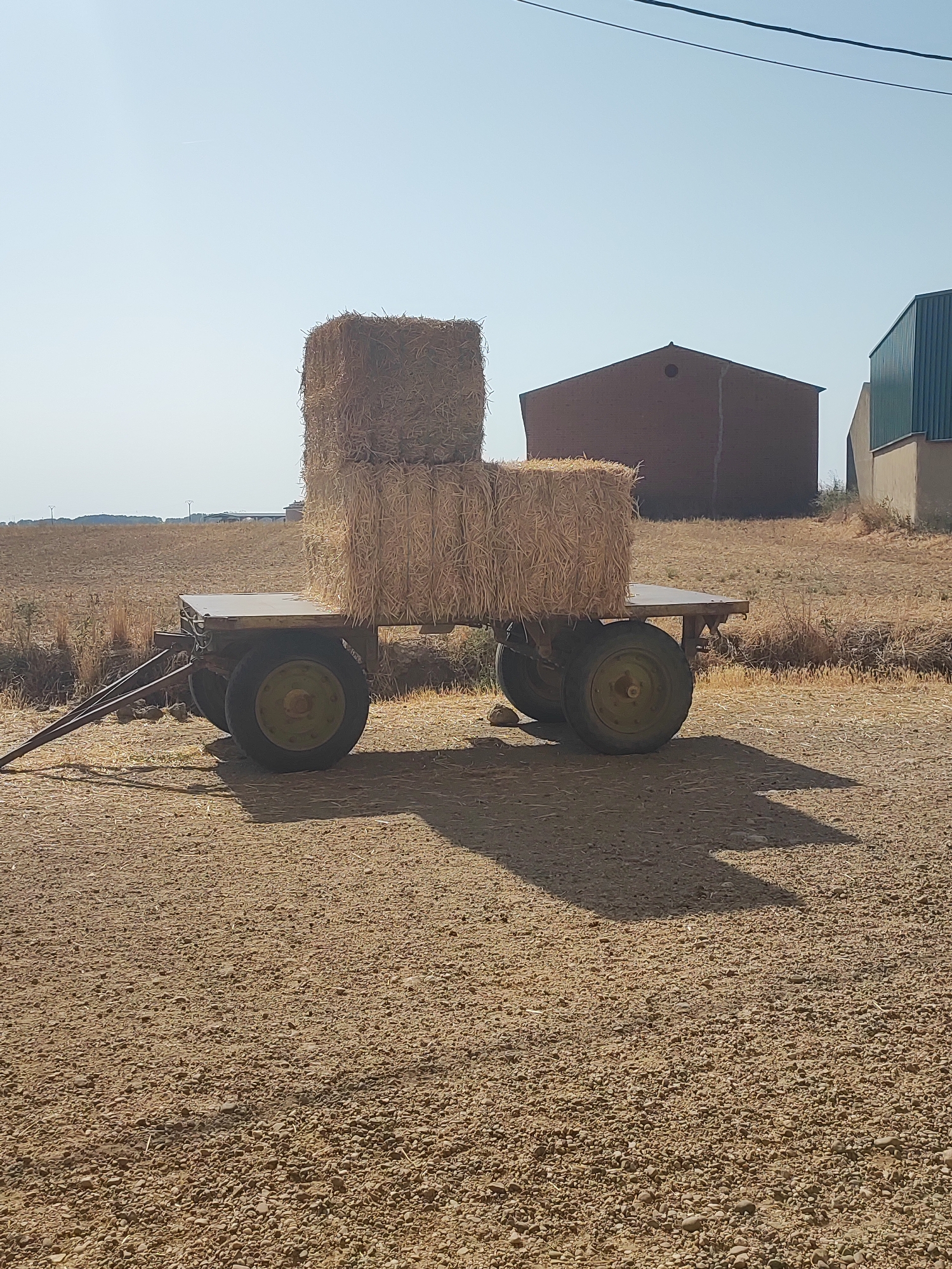A photograph taken on a clear day shows a two-tiered stack of hay bales resting on a flatbed trailer. The trailer has two axles, each with two wheels. The trailer is positioned on a dirt ground, with dry, mowed grass visible in the background. In the distance, there are several buildings, including a reddish-brown barn-like structure and a taller, greenish-blue building. The sky is a pale blue with a faint line that could be a contrail. The scene appears to be in a rural area, possibly a farm or agricultural field. The location is identified as Autillo De Campos, Spain. There are no people or animals visible in the image. The lighting suggests it is daytime, with shadows cast by the trailer and hay bales indicating the sun is to the left of the frame.