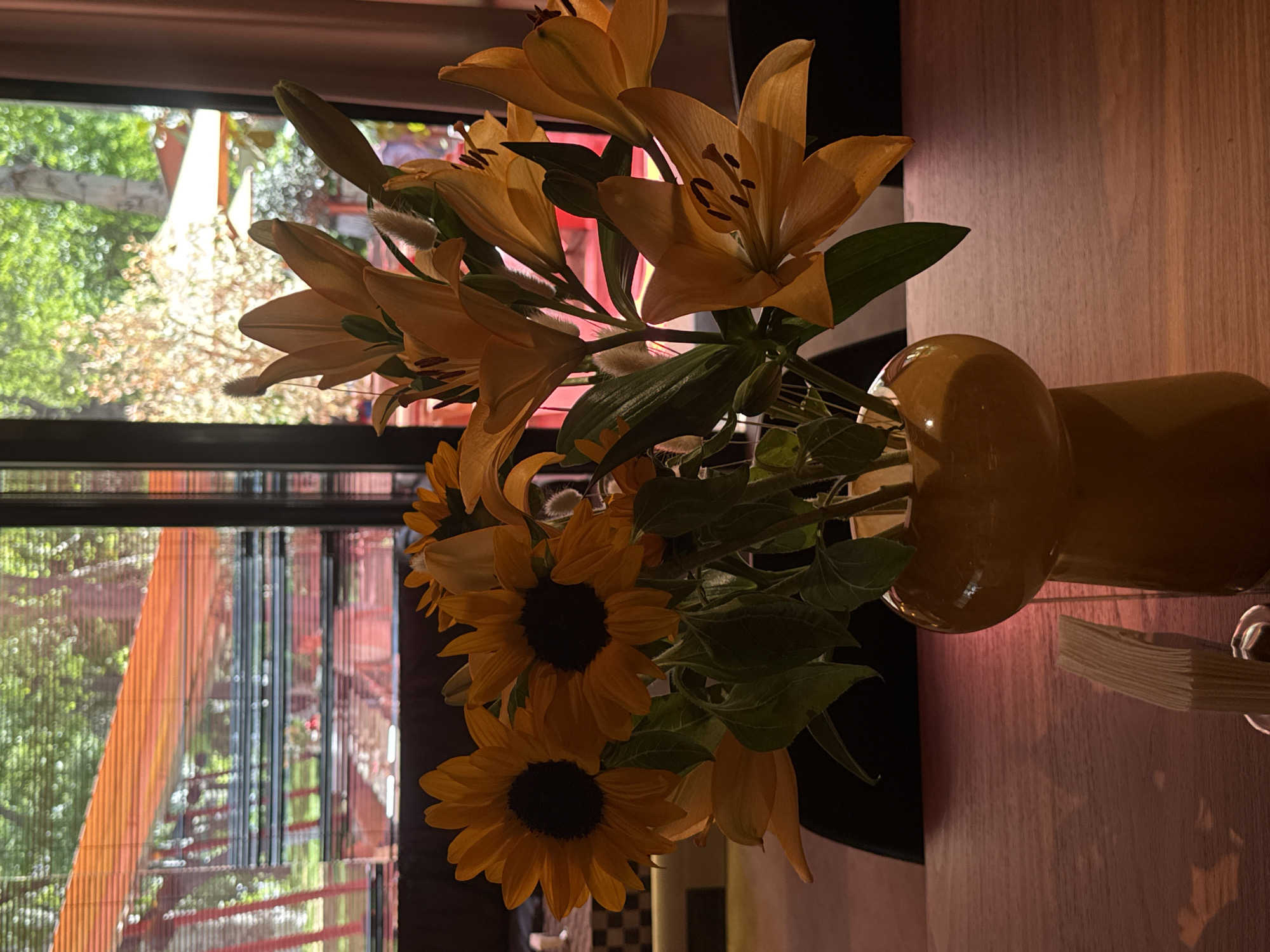 A still life of yellow lilies and sunflowers arranged in a yellow vase sits on a wooden table against a backdrop of a window. The window provides a view of a cityscape, potentially located in Yerevan, Armenia, though specific landmarks are indistinct. The city view through the window is slightly blurred due to focus and possibly distance, showing elements of buildings and greenery. The vase is a distinctive rounded shape. Adjacent to the vase on the table is a stack of papers or cards. Lighting is a mix of natural light from the window and artificial light, creating shadows and highlights on the flowers and vase.