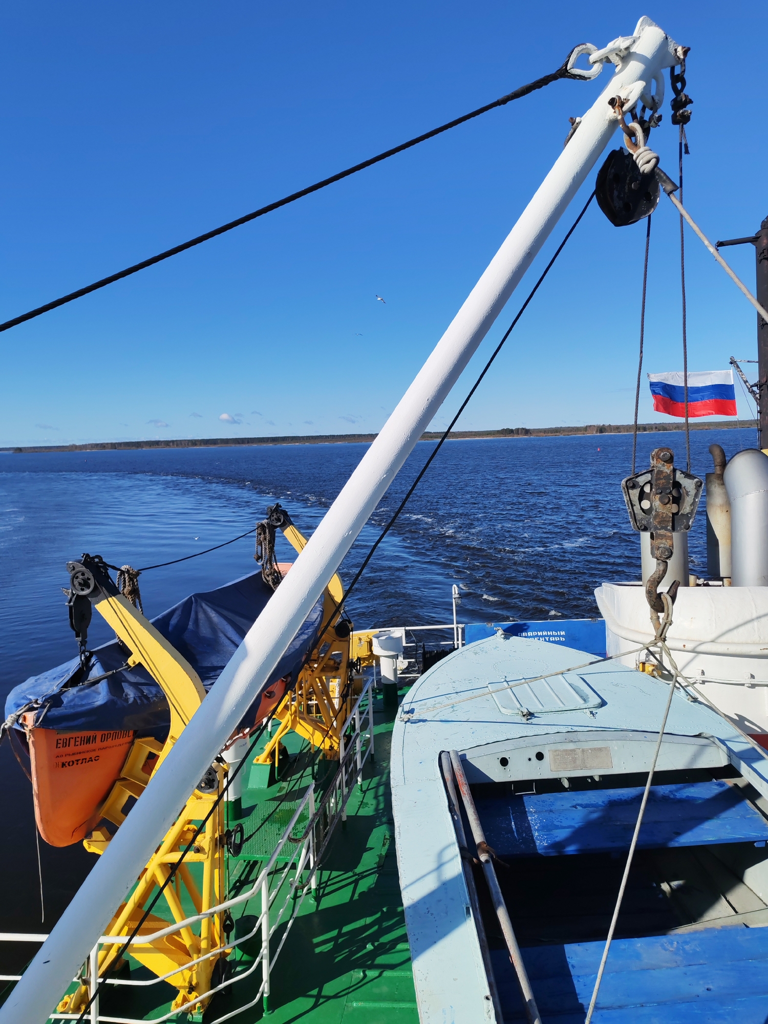 The image depicts the deck of a boat sailing on a body of water, likely a river or lake. The water appears blue and calm with visible wake trails. Along the horizon, a line of trees marks the distant shoreline.
The foreground features various elements of the boat's deck. A light blue lifeboat sits secured on the deck. Nearby, an orange lifeboat is suspended by a yellow davit. A white, angled boom with rigging and pulleys is prominent in the view.
A Russian flag is visible in the upper right of the frame, attached to a pole. The sky above is a clear blue with a few visible clouds and a bird flying.