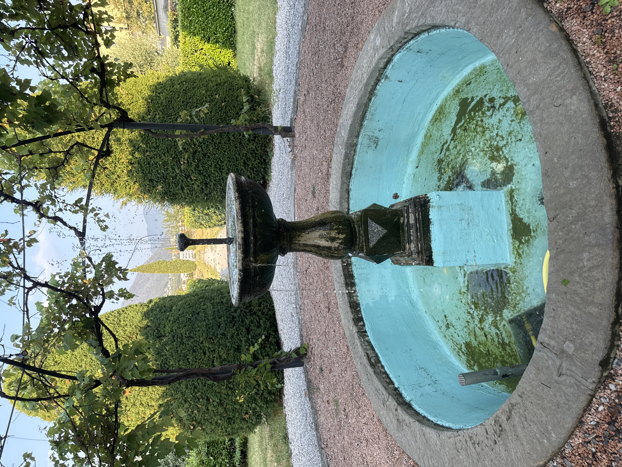 The image shows an outdoor fountain in a garden setting. The fountain is made of stone and has a tiered design, with water spouting from the top. The basin of the fountain is painted turquoise and contains some green algae. In the background, there are manicured green hedges, trees, and a gravel path. The scene appears to be in a temperate climate, possibly during the daytime, with natural lighting. The overall impression is one of a well-maintained garden with a central water feature. There are no people or animals visible in the image. The context of Balerna, Switzerland, suggests a European garden style.