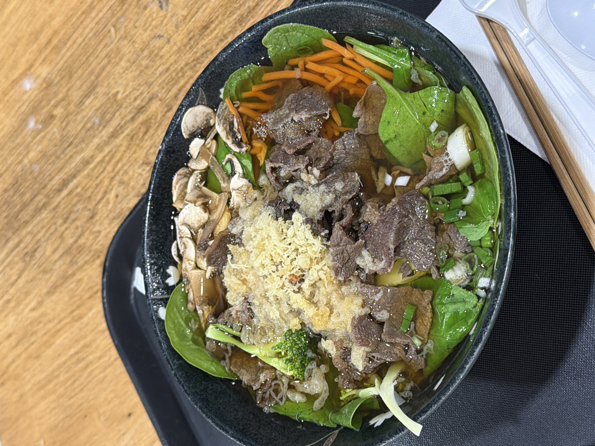 The image shows a bowl of beef pho, a Vietnamese noodle soup, on a table. The soup contains sliced beef, shredded carrots, spinach, mushrooms, broccoli, scallions, and possibly some crispy fried shallots. The bowl is black and sits on a black tray. Next to the bowl are chopsticks, a spoon, and a fork. The table appears to be made of wood. Given the information provided, it is likely the photo was taken at a restaurant or food establishment in Niles, United States of America. There are no people visible in the image.