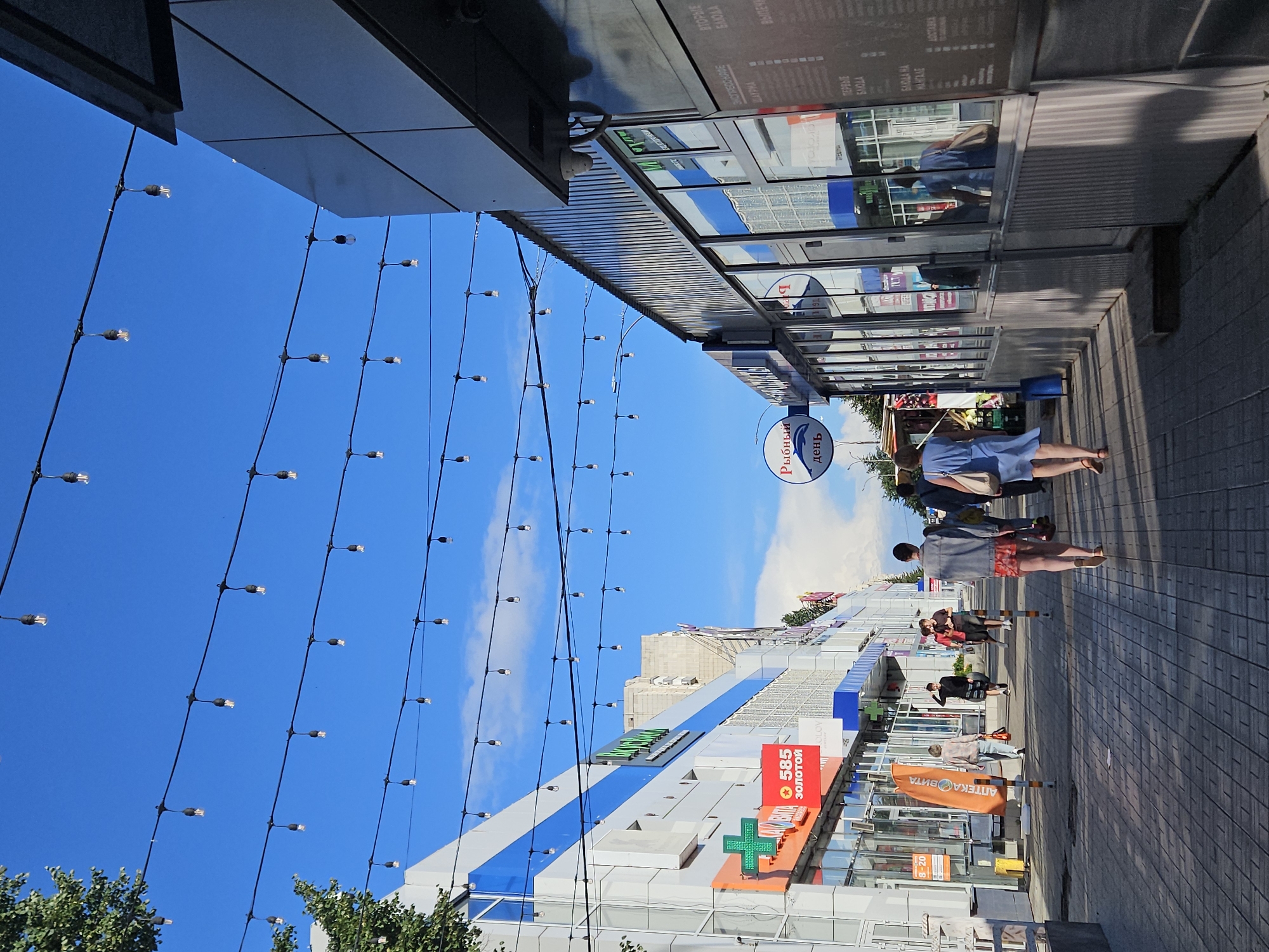 The image shows a street scene in Ulyanovsk, Russia, with a pedestrian walkway lined with shops. A string of decorative lights hangs overhead against a bright blue sky with scattered clouds. Several people are walking along the sidewalk, some alone and some in pairs. On the left side of the street, there are modern buildings with glass storefronts, including a pharmacy with a green cross logo and a store displaying "585 ЗОЛОТОЙ" (585 GOLD) in a red rectangular sign. On the right side, there is a building with a corrugated metal facade and large glass windows, reflecting the surroundings. One shop appears to be a fish market with a circular sign depicting a dolphin and the text "Рыбный день" (Fish Day). The ground is paved with gray rectangular tiles. The overall atmosphere suggests a sunny day in a commercial area.