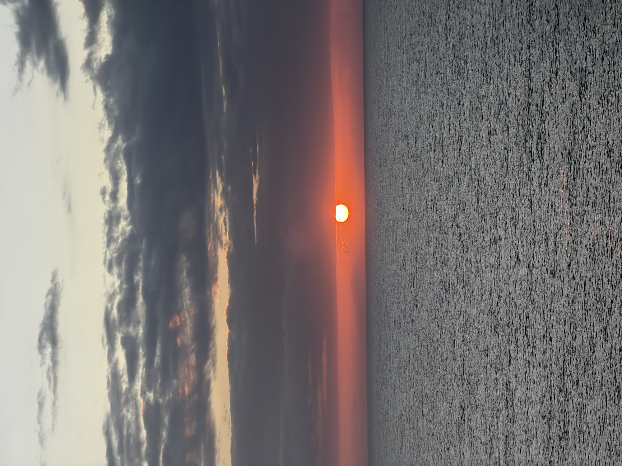 The media file captures a serene sunset or sunrise over a vast body of water. The image is composed with a distinct horizon line separating the sky from the water.
In the upper portion of the image, the sky displays a gradient of colors. A bright, circular sun, glowing intensely in orange and yellow hues, is positioned low on the horizon, slightly to the left of the center. The area of the sky immediately surrounding the sun is illuminated with warm orange and red tones, transitioning upwards into darker shades of blue-grey and muted purples. Numerous dark grey clouds are scattered across the sky, some dense and undulating, with their edges faintly highlighted by the sun's warm light.
The lower half of the image features a wide expanse of water, appearing as a uniform dark grey surface. The water is textured with countless small ripples or subtle waves, creating a consistent pattern across its entirety. There are no visible people, objects, or structures on the water or in the sky, emphasizing the natural and untouched landscape. The time of day is clearly dawn or dusk, indicated by the low position of the sun and the rich, transitional colors of the sky. The weather appears clear over the horizon where the sun is visible, despite the presence of clouds higher up. No visible text is present in the image.