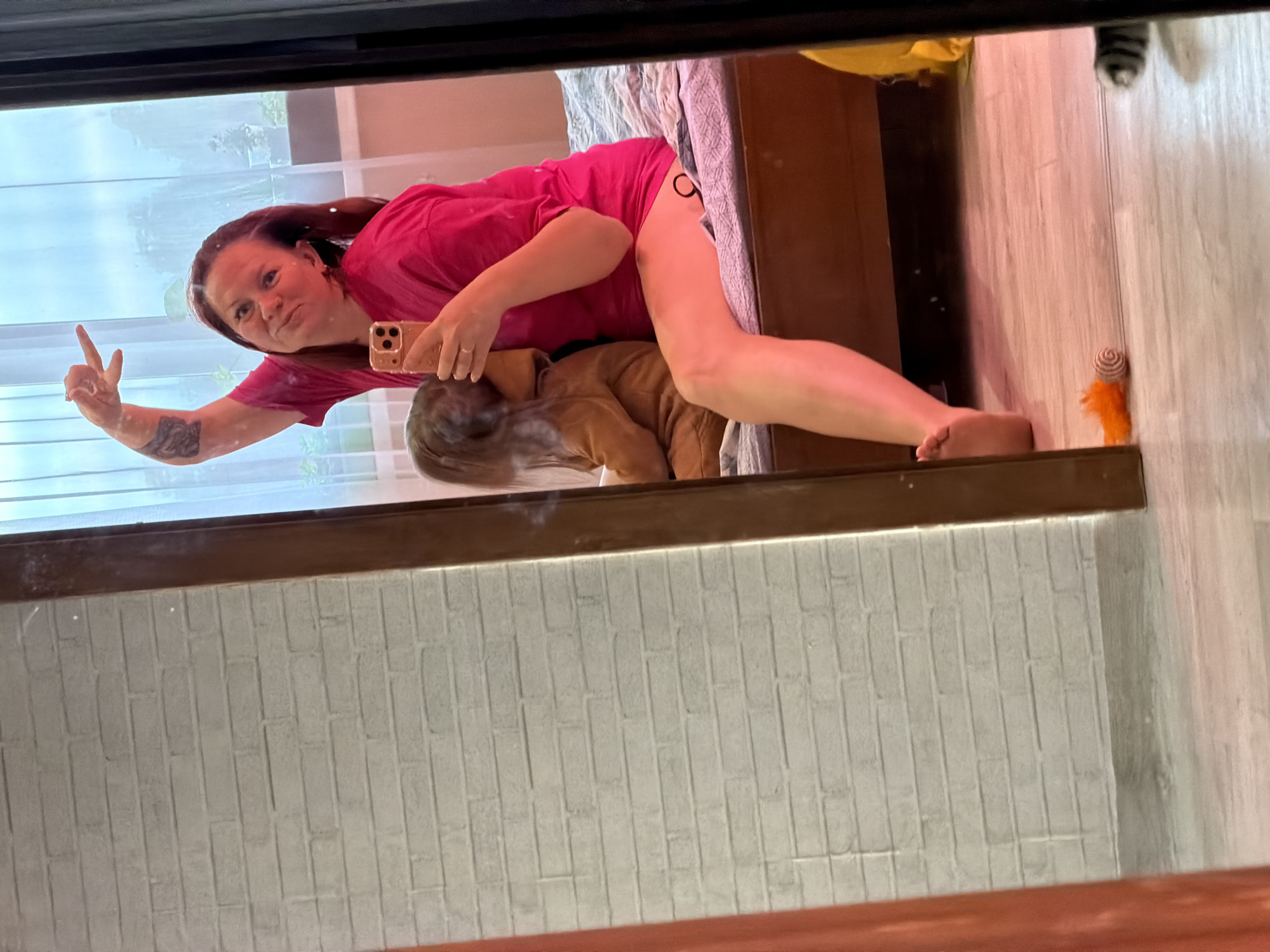 This indoor mirror selfie, taken in Chaikovsky, Russia, features a woman and a child. The woman, with long dark hair and wearing a bright pink t-shirt, is leaning over, likely on a bed with patterned bedding. She is holding a gold-colored smartphone to take the photo and making a peace sign with her left hand. A small tattoo is visible on her left upper arm, and another dark tattoo is on her upper left thigh. She has a faint smile.
Partially hidden beneath the woman is a child, visible from the back of the head and upper body, wearing a brown or tan hooded outfit. The room has a light-colored wooden floor. An orange feathery toy and a striped cat toy are visible on the floor to the right. The mirror's reflection also shows a window with a bright outdoor view of the sky and some greenery, suggesting it is daytime. The lower part of the reflection reveals a wall with a white brick or tile pattern. No text is visible in the image.