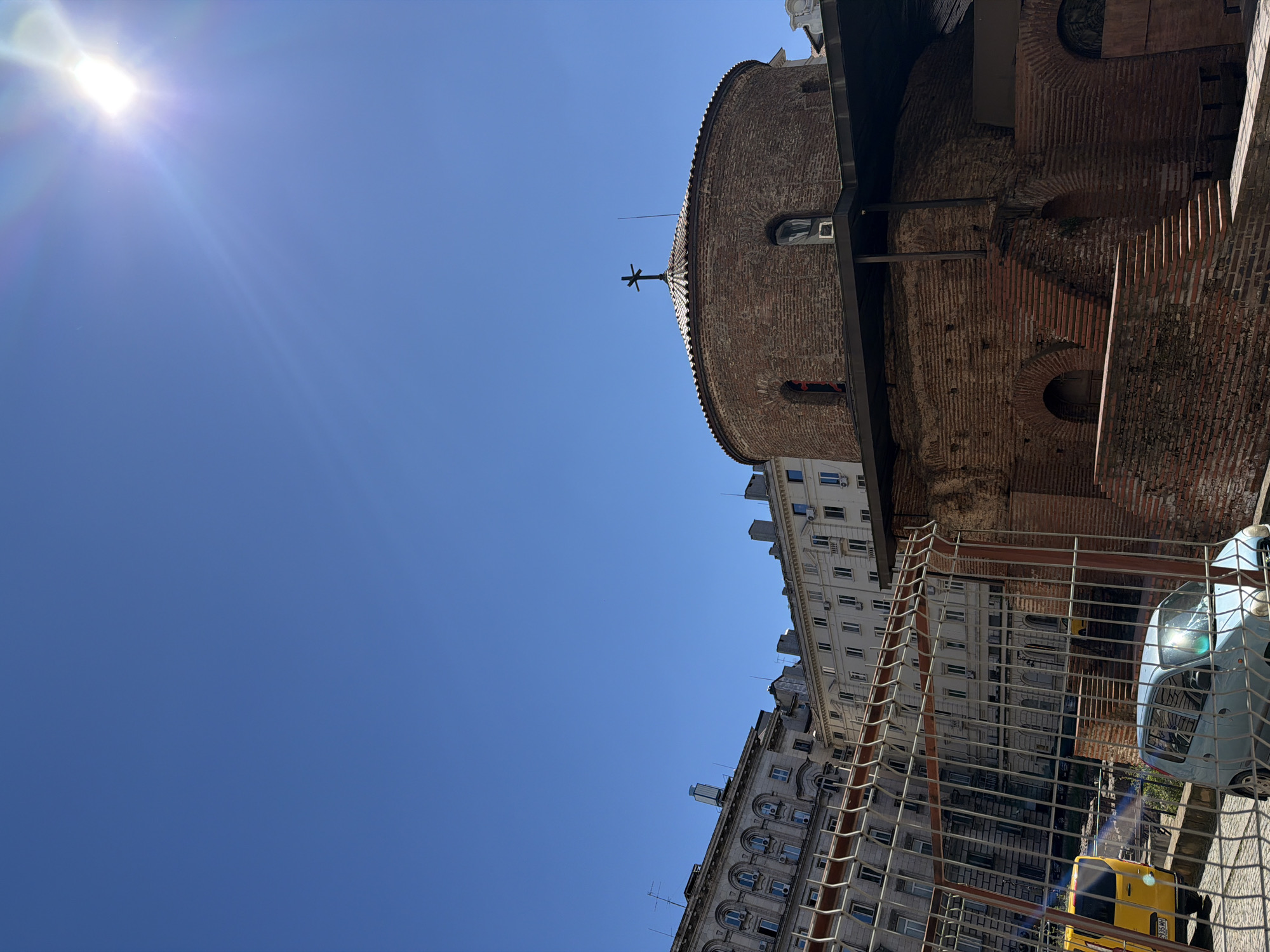 The image shows a low-angle view of the Church of St. George Rotunda in Sofia, Bulgaria, set against a bright blue sky with the sun in the upper left corner. The church is constructed of red brick, with a rounded top and a cross on its apex. To the right of the church are the facades of adjacent buildings. Construction barriers are in the foreground. The scene appears to be taken during a sunny day in an urban environment.