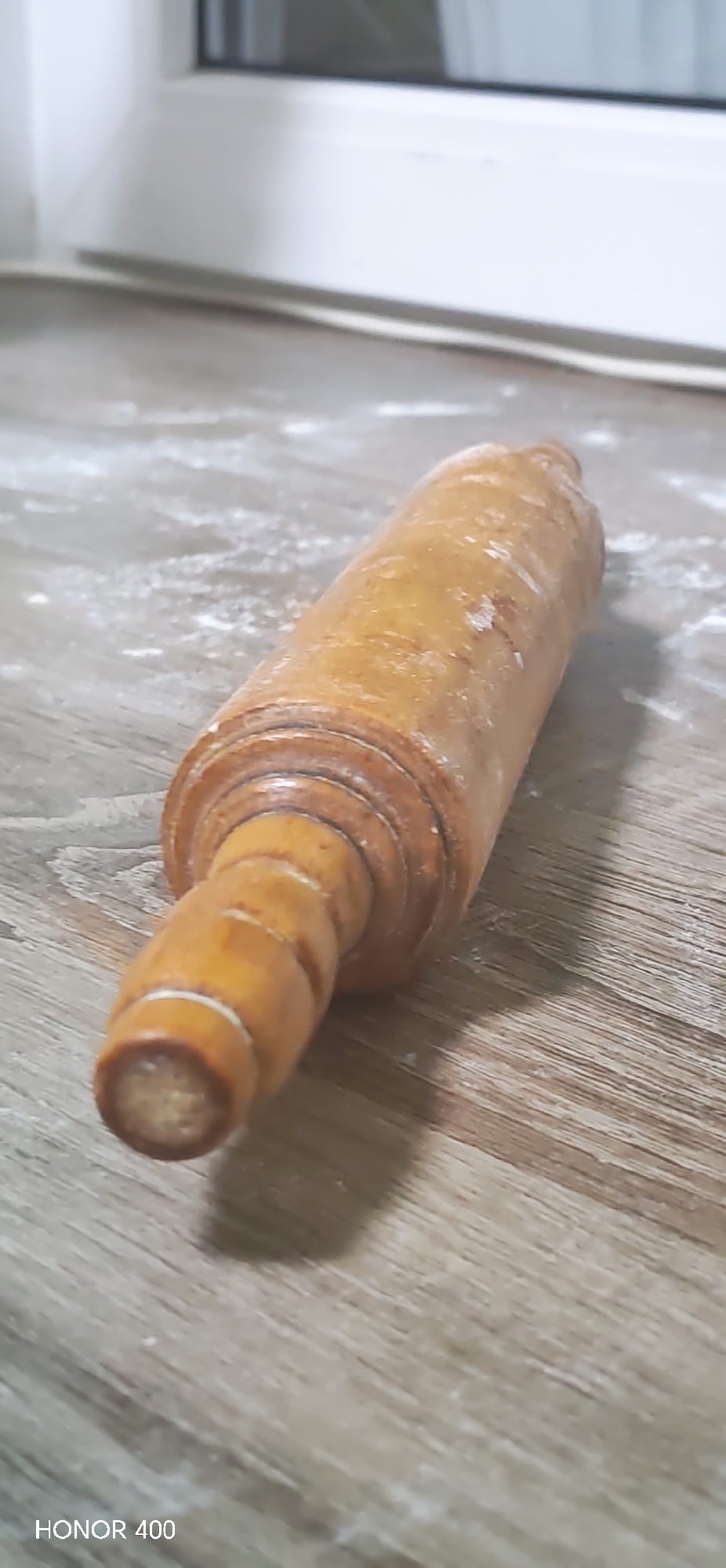 A wooden rolling pin rests on a surface, possibly a countertop, lightly dusted with flour. The surface has a wood grain pattern. A window is visible in the background. "HONOR 400" is displayed in the lower left corner of the image. There is no indication of people or any action in the image. The location within Noviy, Russia, cannot be determined from the visual information provided.