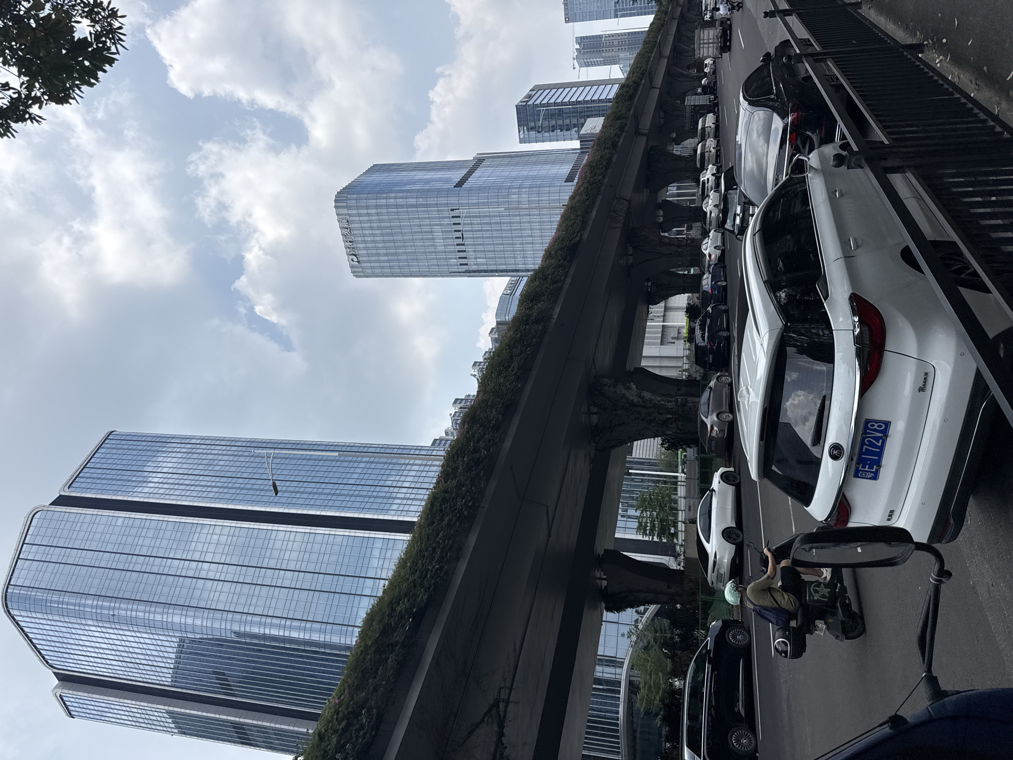 This vertical photograph captures an urban landscape in Huangpu District, China, under a largely overcast sky with patches of brighter blue. Towering modern skyscrapers with reflective glass facades dominate the background and mid-ground. One prominent building in the center displays large Chinese characters, which appear to be "平安银行" (Ping An Bank). Another similar skyscraper is visible on the far left.
An elevated concrete road or overpass stretches horizontally across the middle of the frame, extensively covered with dense green foliage, suggesting an effort at urban greening. Beneath this structure, a street is lined with numerous parked vehicles. In the foreground, a white SUV with the Chinese license plate "粤E·172V8" is clearly visible, confirming the Guangdong province location. Small text "TRiMAX" is also discernible on its rear bumper. To the right of the white car, a person wearing a light blue helmet and a backpack is seated on a black scooter or motorcycle, positioned on the road. A dark tree with lush leaves is partially visible in the upper left corner.
The scene portrays a typical daytime city environment, characterized by modern architecture, bustling street activity, and urban infrastructure.