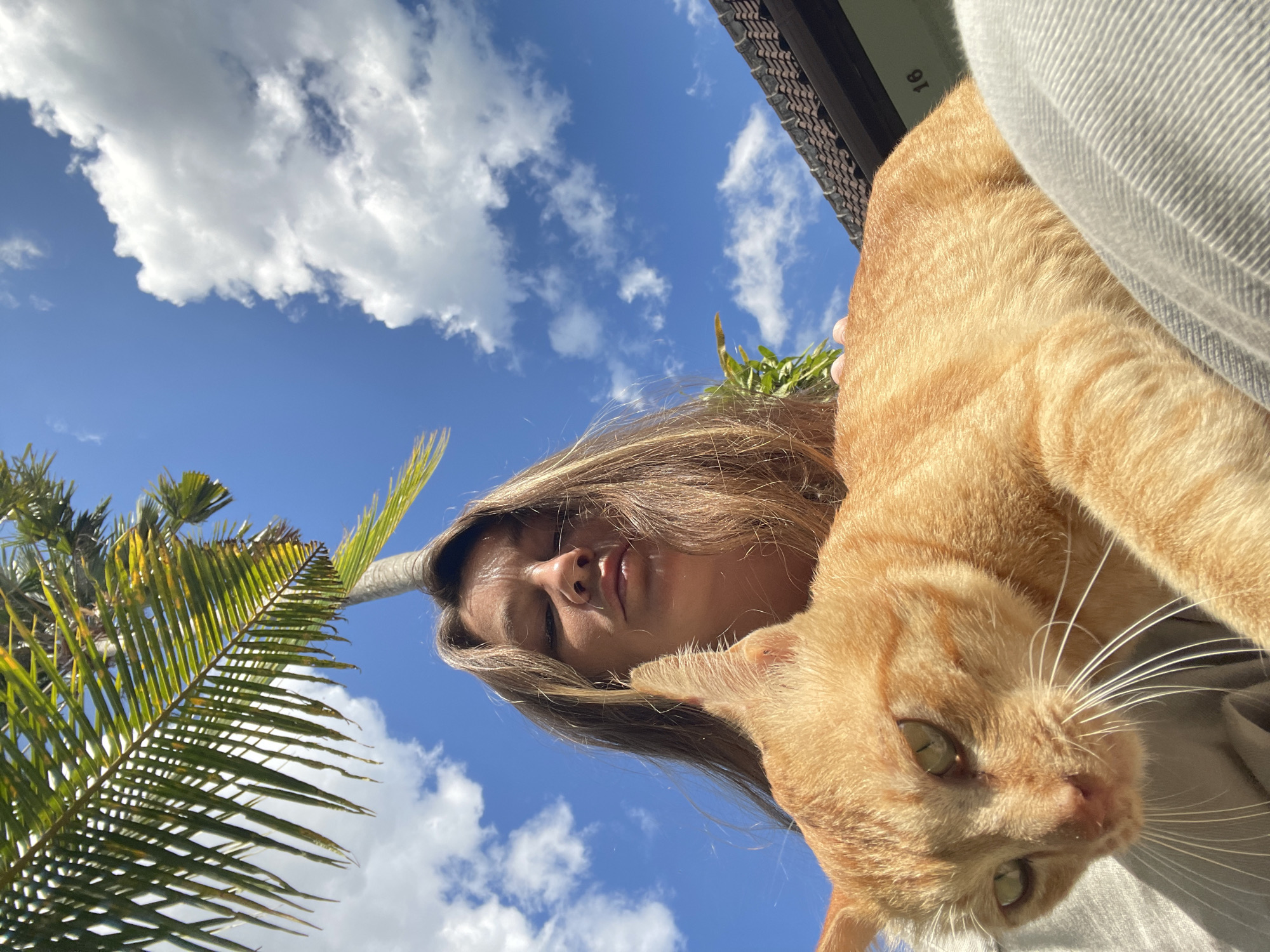 A low-angle, close-up, outdoor, waist-up selfie shows a young woman with long, light brown hair holding an orange tabby cat. The woman's face is partially obscured by her hair and the cat, but her eyes are visible and directed downward. The cat, positioned in the foreground, has green eyes and is looking towards the viewer. The woman is wearing a light-colored top and light-colored pants. Behind them, a clear blue sky with scattered white clouds is visible, along with a palm tree frond in the upper left corner and a tiled roof of a building in the upper right corner. The scene appears to be in North Kuta, Indonesia, given the tropical vegetation and architecture.