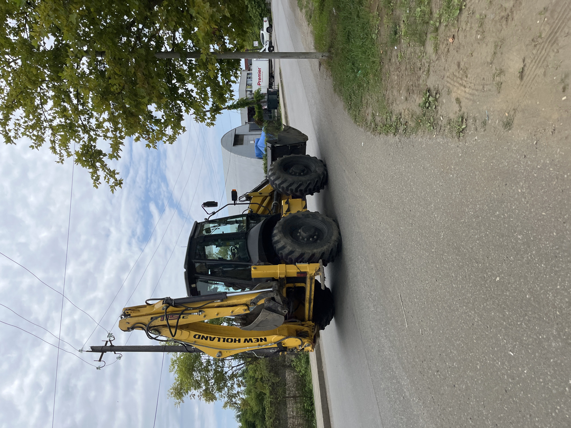 A yellow New Holland backhoe loader is parked on a paved road in Ureki, Georgia. The backhoe is positioned at an angle, facing slightly away from the camera. Its bucket is lowered, and the boom is extended upwards. The cab of the backhoe is dark and reflective. In the background, there is a long, light-colored building with a curved roof. A white truck with "Premier" written on its side is parked near the building. To the right of the backhoe, there is a green tree with large leaves, and a utility pole with electrical wires runs diagonally across the sky. The sky is partly cloudy. The road appears to be a two-lane road, with grass and dirt on the side. The overall scene suggests a construction or industrial area on the outskirts of the town.