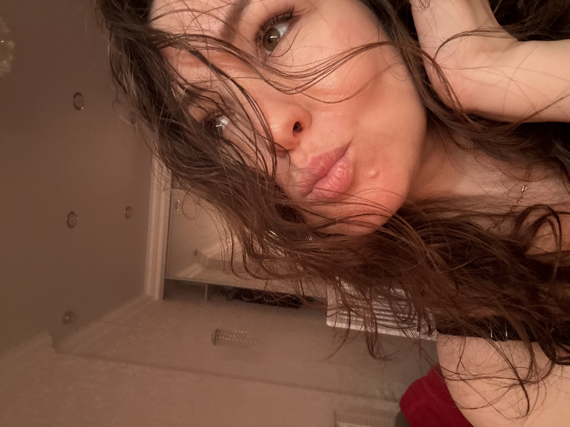 A close-up selfie shows a woman with dark, wet-looking hair framing her face. Her head is tilted, and she looks directly at the camera with brown/hazel eyes. Her lips are slightly pursed in a "kissy" expression, and a small blemish is visible on her right cheek. Part of her arm or hand is raised near her head. She wears a gold-colored chain or necklace. In the background, behind her, a light-colored wall with several circular indentations, suggestive of jacuzzi jets, is visible. Below, a reflective surface, possibly water or wet tiles, is present. The indoor lighting is warm and somewhat dim. A dark garment or strap is seen near her shoulder, and a piece of red fabric, possibly a towel, is visible at the bottom right. No text is visible in the image.