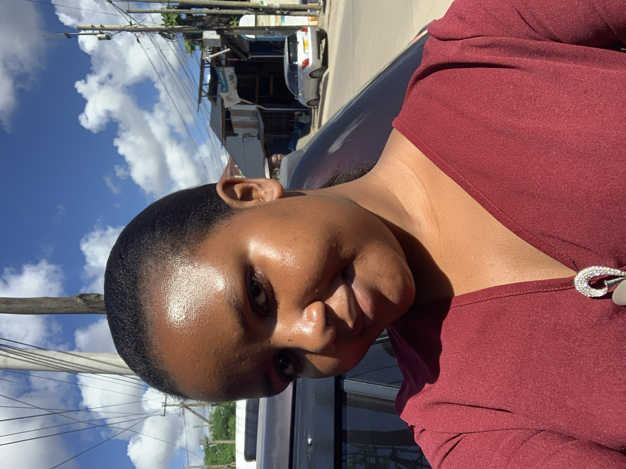 A close-up, tilted shot shows a dark-skinned woman with short, dark hair standing next to a dark-colored car. She is wearing a maroon top with a decorative pin on the neckline. The background includes a street scene with power lines, buildings, and a white car. The location is Dar es-Salaam, Tanzania.