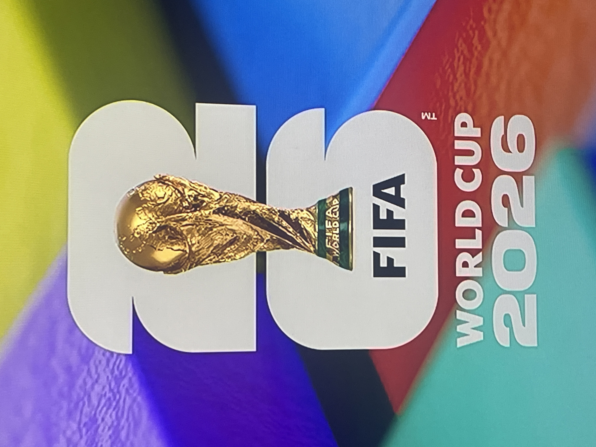 This is an image of the FIFA World Cup 2026 logo. The logo features the FIFA World Cup trophy prominently in the center, superimposed over large, stylized numbers "2" and "6" which represent the year 2026. Below the trophy and numbers, the text "FIFA WORLD CUP 2026" is displayed. The background is a vibrant, abstract design with diagonal stripes of various colors, including purple, yellow, blue, red, and green. There is no indication of the city of Ruse, Bulgaria, or any specific location within it. No people or specific actions are depicted in the image.