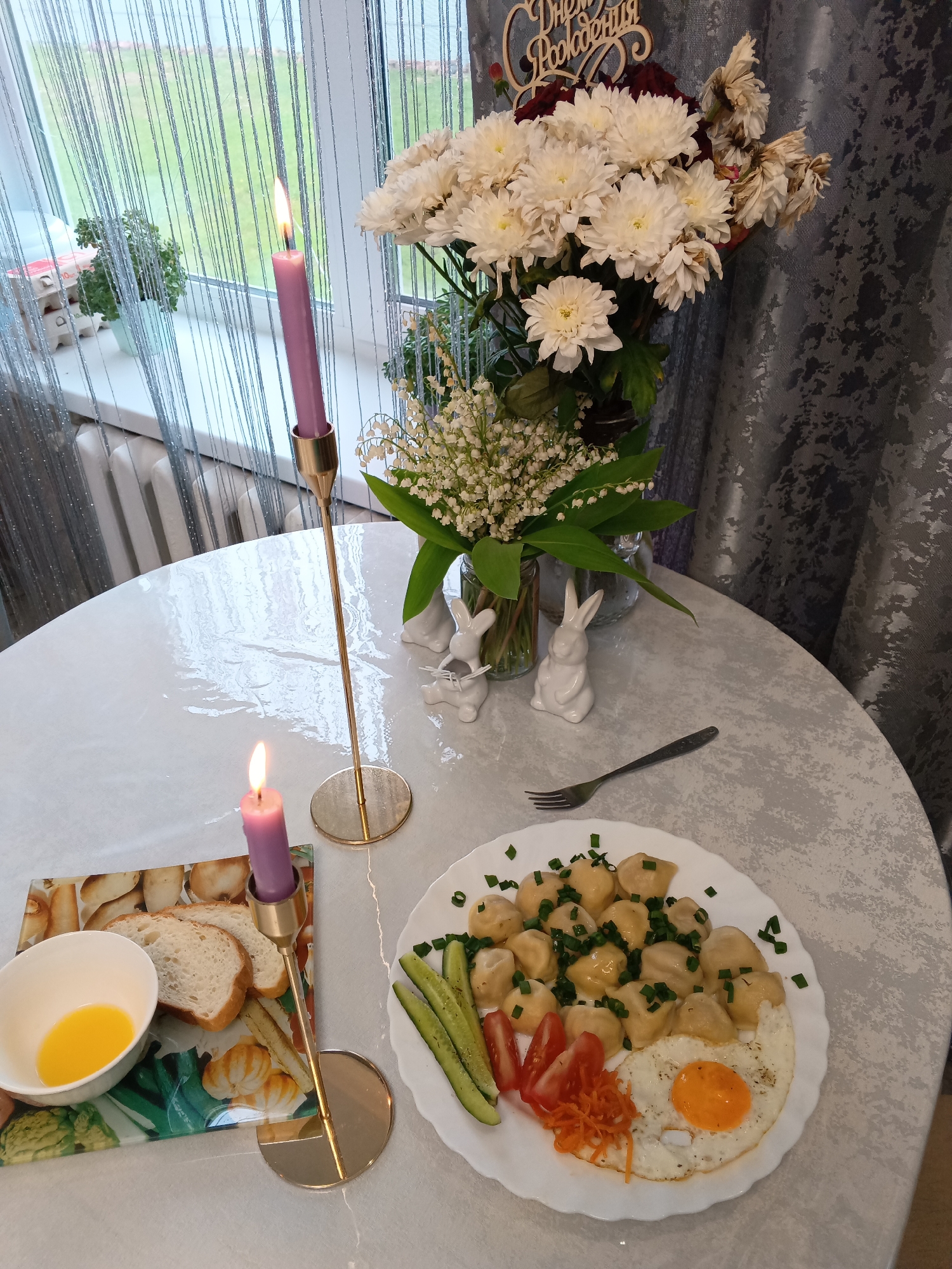 The image shows a table setting in what appears to be a domestic environment, likely a home in Andreyevo, Russia. The table, covered with a light-colored tablecloth with a subtle pattern, is set with a meal of dumplings, garnished with chopped green onions, and an over-easy egg, along with sliced cucumbers, tomatoes, and shredded carrots on a white plate. A fork is placed beside the plate. To the left, there is a cutting board with sliced bread and a small bowl containing a yellow liquid, possibly melted butter or egg yolk.
Two tall, brass candle holders are present, each holding a lit purple candle. In the background, there is a vase filled with white chrysanthemums and lily of the valley flowers. Two small white ceramic rabbit figurines sit near the vase. A decorative wooden sign with Cyrillic text, possibly a birthday greeting, is placed among the flowers.
The background features a window with light-colored curtains, revealing glimpses of the exterior, including greenery. The room appears to be illuminated by both natural light from the window and the candlelight. The overall scene suggests a celebration or a special meal arrangement.