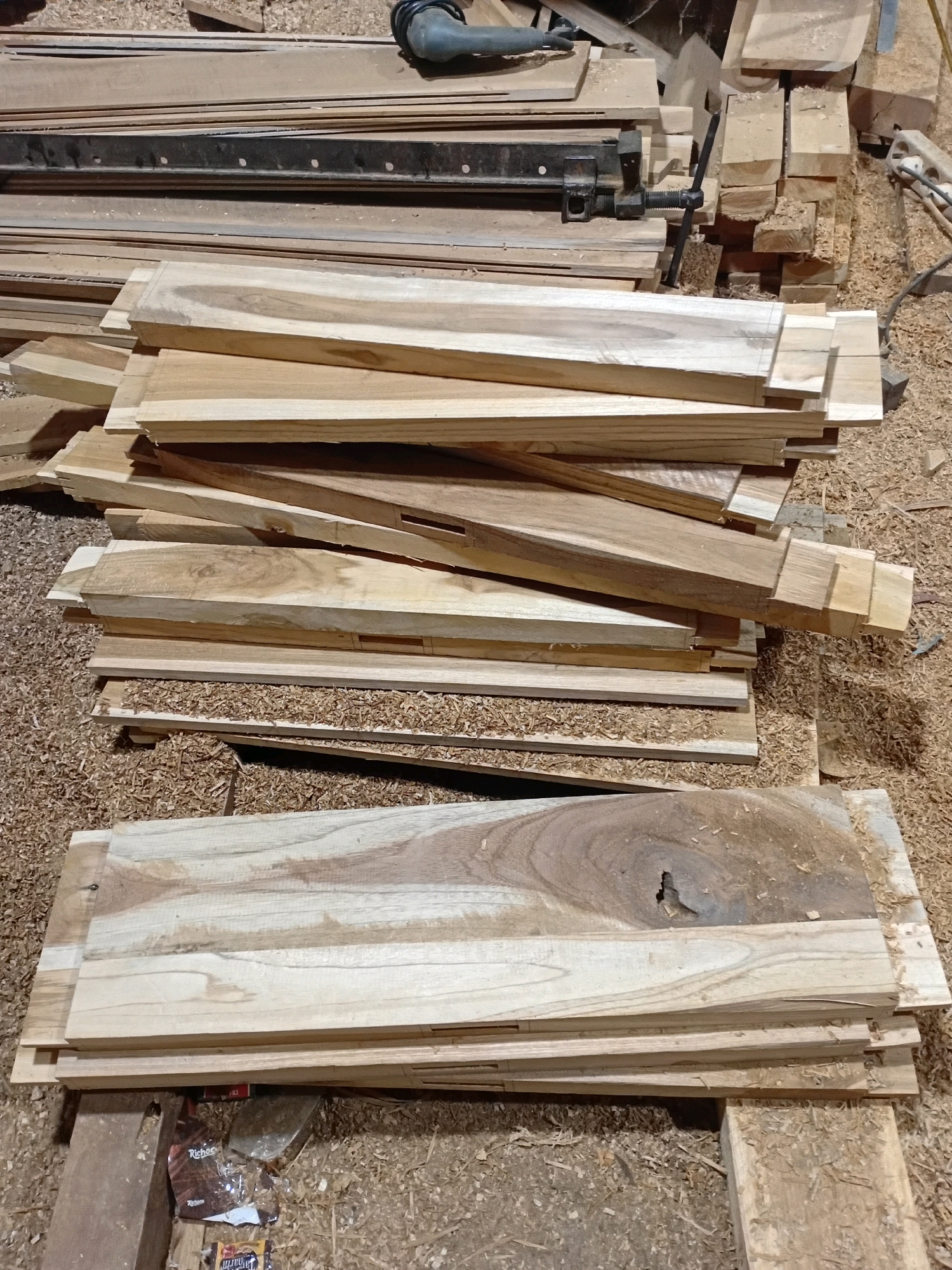 A stack of rough-cut wooden planks is piled on a surface covered in wood shavings. The planks vary in size and color, with some showing distinct grain patterns and knots. In the background, more lumber and woodworking tools, including what appears to be a clamp and a power tool, are visible. The scene suggests a workshop or lumberyard environment. The location is Playen, Indonesia.
