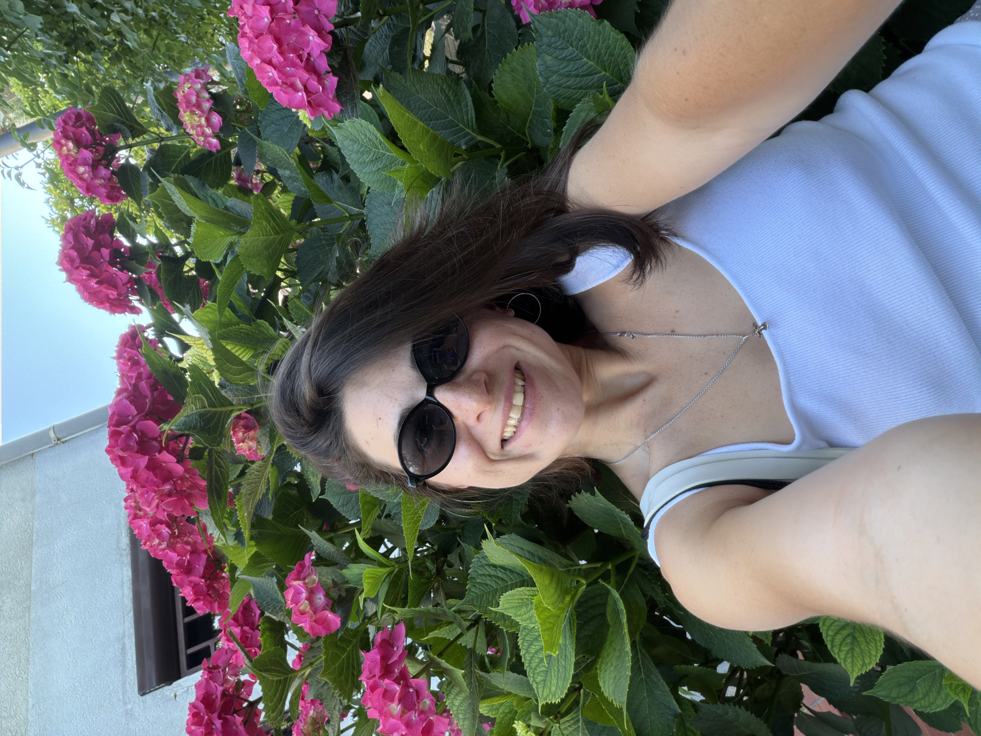 The image shows a woman with brown hair, wearing sunglasses and a white top, taking a selfie in front of a bush with pink hydrangeas. The background includes a building with a window. The location appears to be outdoors, possibly in Budva, Montenegro, given the presence of hydrangeas, which are common in the region.