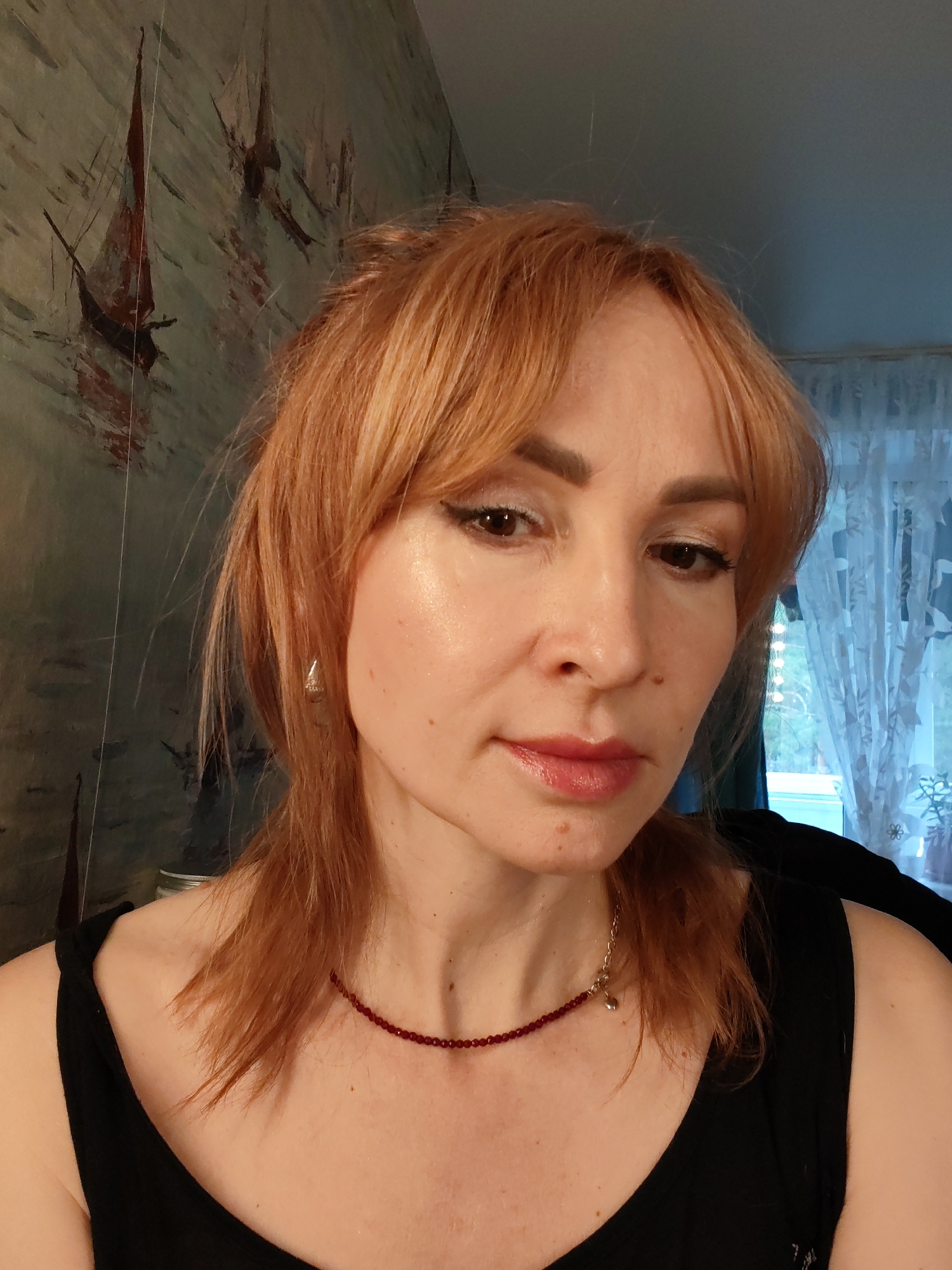 A middle-aged woman with reddish-blonde hair and light skin is facing the camera. She has brown eyes and is wearing makeup, including eyeliner and lipstick. She is wearing a black tank top and a red beaded necklace. A silver earring is visible in her left ear.
Behind her, there is a wall with a painting depicting boats, and to the right, a window with sheer curtains. The location is likely an indoor space, possibly a home. The available information does not confirm that the image was taken in the city of Chaikovsky, Russia.