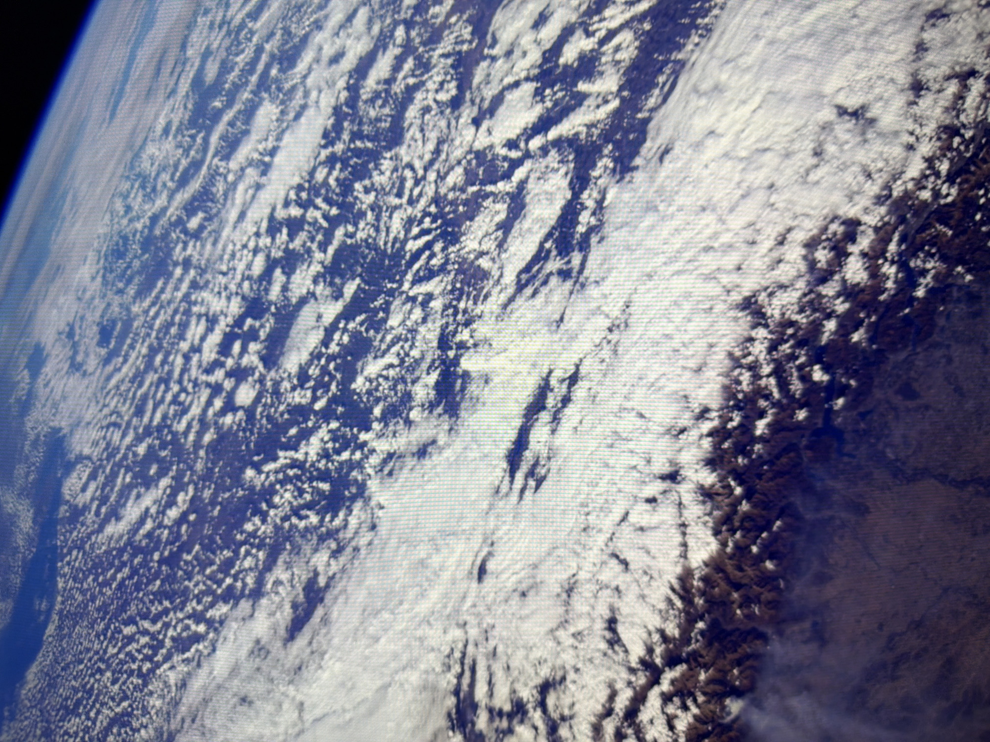 This is an aerial photograph of Earth's atmosphere and surface, taken from space. The image displays a vast expanse of clouds, appearing as a textured white and gray blanket, obscuring much of the land below. The clouds exhibit patterns of turbulent formations, suggesting weather systems. Below the clouds, rugged, dark brown terrain is visible, characterized by sharp ridges and peaks, indicative of a mountainous region. The overall impression is a high-altitude view of a cloudy planet with visible mountainous topography. The image does not depict the city of Kyiv, Ukraine.