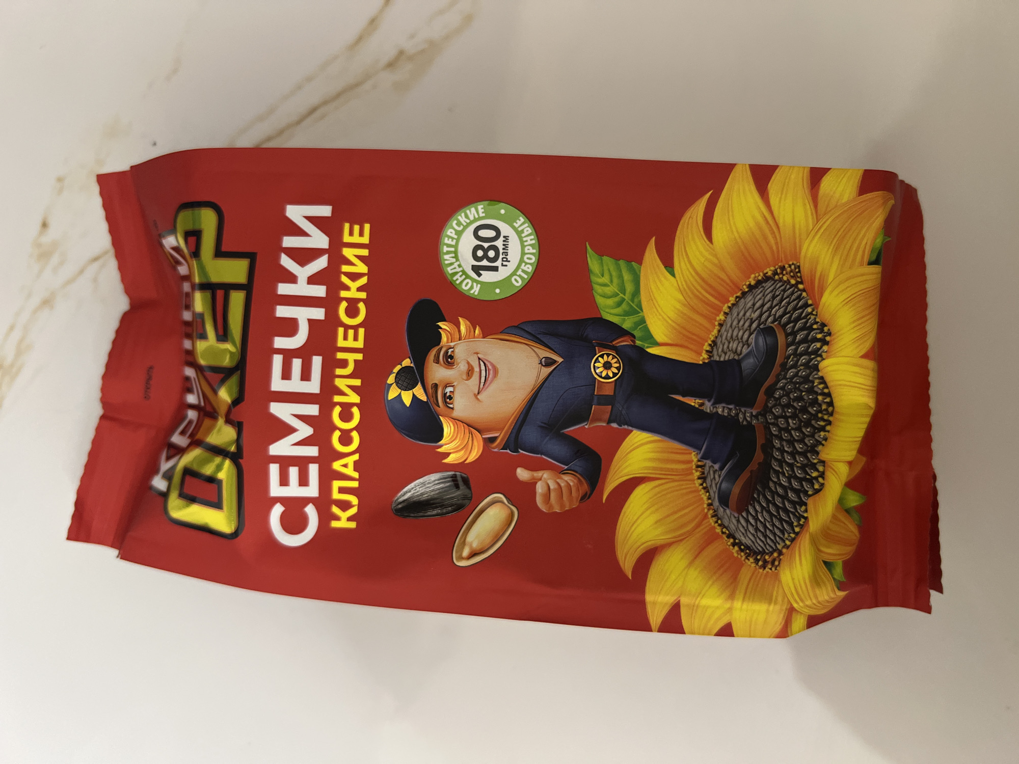 A red, flexible pouch-style package for sunflower seeds is centrally displayed on a multi-surface background. The package features the brand name "ОХЕР" (Okher) in yellow and black stylized letters at the top, followed by "СЕМЕЧКИ" (Sunflower Seeds) in large white capital letters, and "КЛАССИЧЕСКИЕ" (Classic) in yellow capital letters. A cartoon character with a fair complexion, orange hair, a dark blue cap with a sunflower emblem, and a dark blue outfit including a jacket, pants, and boots, stands prominently. The character is giving a thumbs-up with its right hand and is positioned on the dark seed-bearing center of a large, vibrant yellow sunflower, with green leaves visible beneath it. To the character's upper left are depicted a single black-shelled sunflower seed and a single peeled sunflower kernel. To the character's right, a circular green label states "КОНДИТЕРСКИЕ" (Confectionery Grade) at the top, "180 грамм" (180 grams) in the center, and "ОТБОРНЫЕ" (Selected) at the bottom. The top edge of the package shows minor crimping. The package rests on a light-colored surface, with a plain white area on the left and a light-toned marble-patterned area with subtle veining on the right.