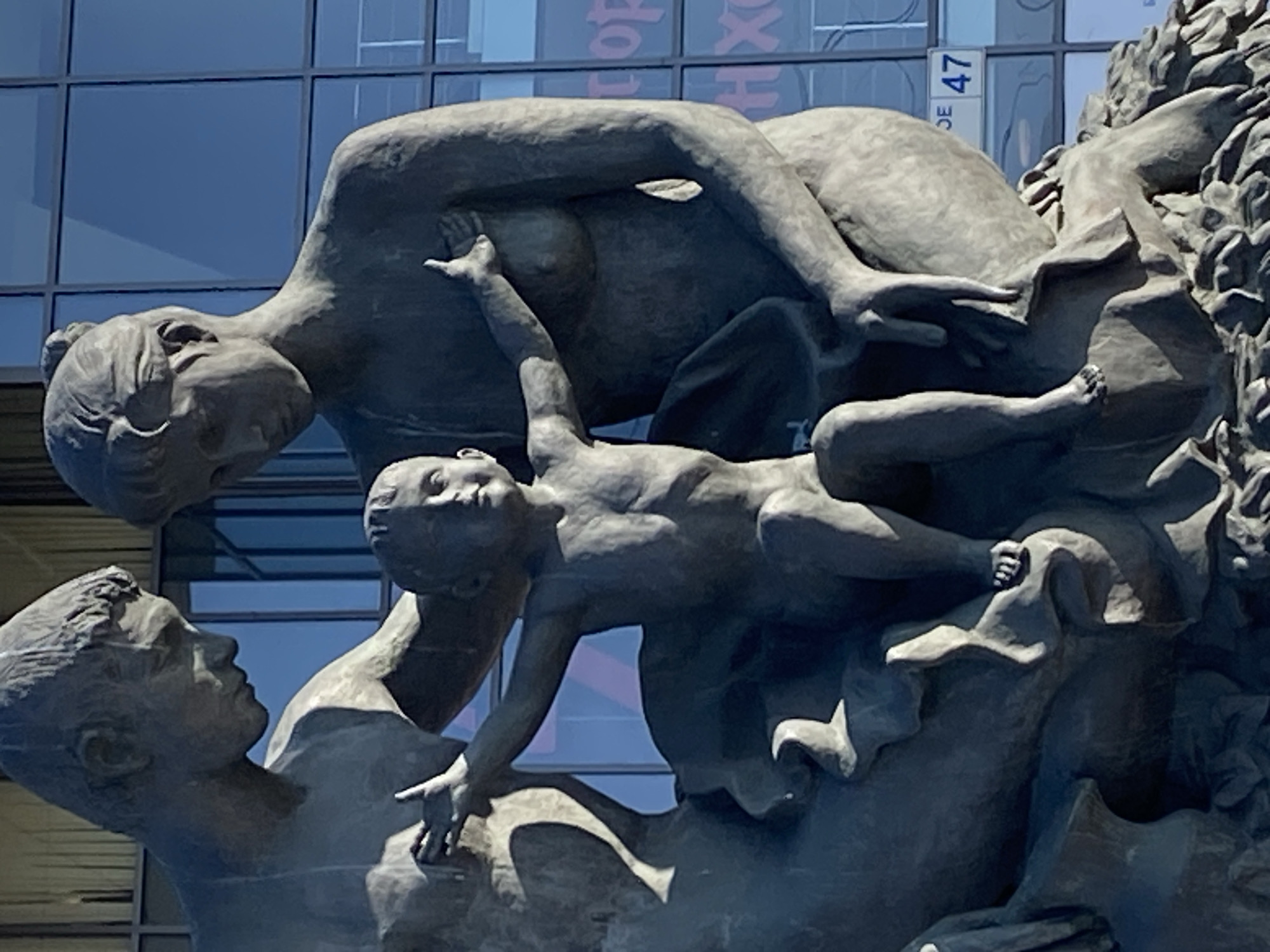 The image shows a close-up of a bronze sculpture depicting a family unit. A man and a woman, sculpted in a neoclassical style, are positioned behind and to the sides of a child who is seated in the center. The child is looking forward with arms outstretched, while the adults are looking down at the child. The man, on the left, has his hand on his chest, and the woman, on the right, also has her hand on her chest. The sculpture is situated outdoors, with a modern building featuring large windows in the background. The windows reflect the sky and parts of the city, including a sign that reads "47" in a square. This sculpture is located at the entrance of the Children's Polyclinic No. 47 on Prospekt Veteranov in Saint Petersburg, Russia.