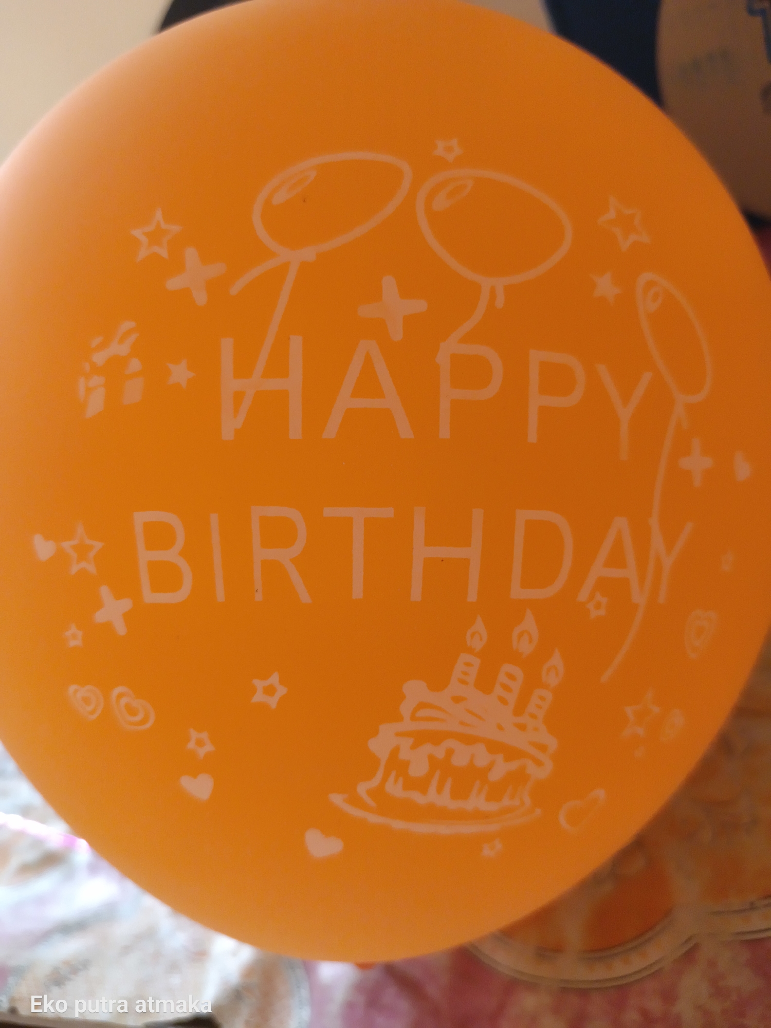 An orange balloon with white lettering and drawings is the central subject of this close-up, eye-level photograph. The balloon is decorated with the words "HAPPY BIRTHDAY" in large white block letters. Surrounding the text are various white drawings, including stick figures holding balloons, stars, hearts, gift boxes, and a birthday cake with lit candles. The balloon is inflated and appears to be resting on a surface that is out of focus and appears to be pink and white patterned fabric. The background is blurred and indistinct. At the bottom left of the image, the text "Eko putra atmaka" is visible in white lettering. The overall impression is that this is a celebratory item, likely for a birthday party. The image does not provide information about a specific location in the city of Pelanjau Jaya, Indonesia.