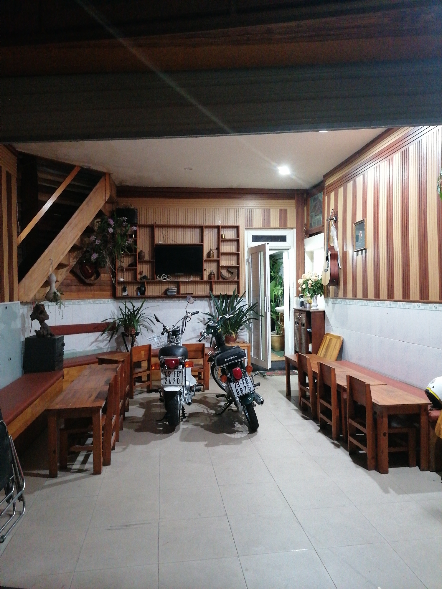 The image depicts the interior of a building, potentially a shop or restaurant, in Da Lat, Vietnam. The space features tiled flooring and walls partially covered with white tiles and partially with brown and tan wood paneling. There are wooden tables and benches arranged along both sides of the room. Two motorcycles are parked in the middle of the room. A staircase is visible on the left side. In the back wall, there is a television mounted on a wooden shelf. An open doorway leads to an area with greenery and possibly an outdoor space. The lighting appears to be a combination of natural and artificial light sources. The overall impression is a functional, somewhat cluttered interior space.