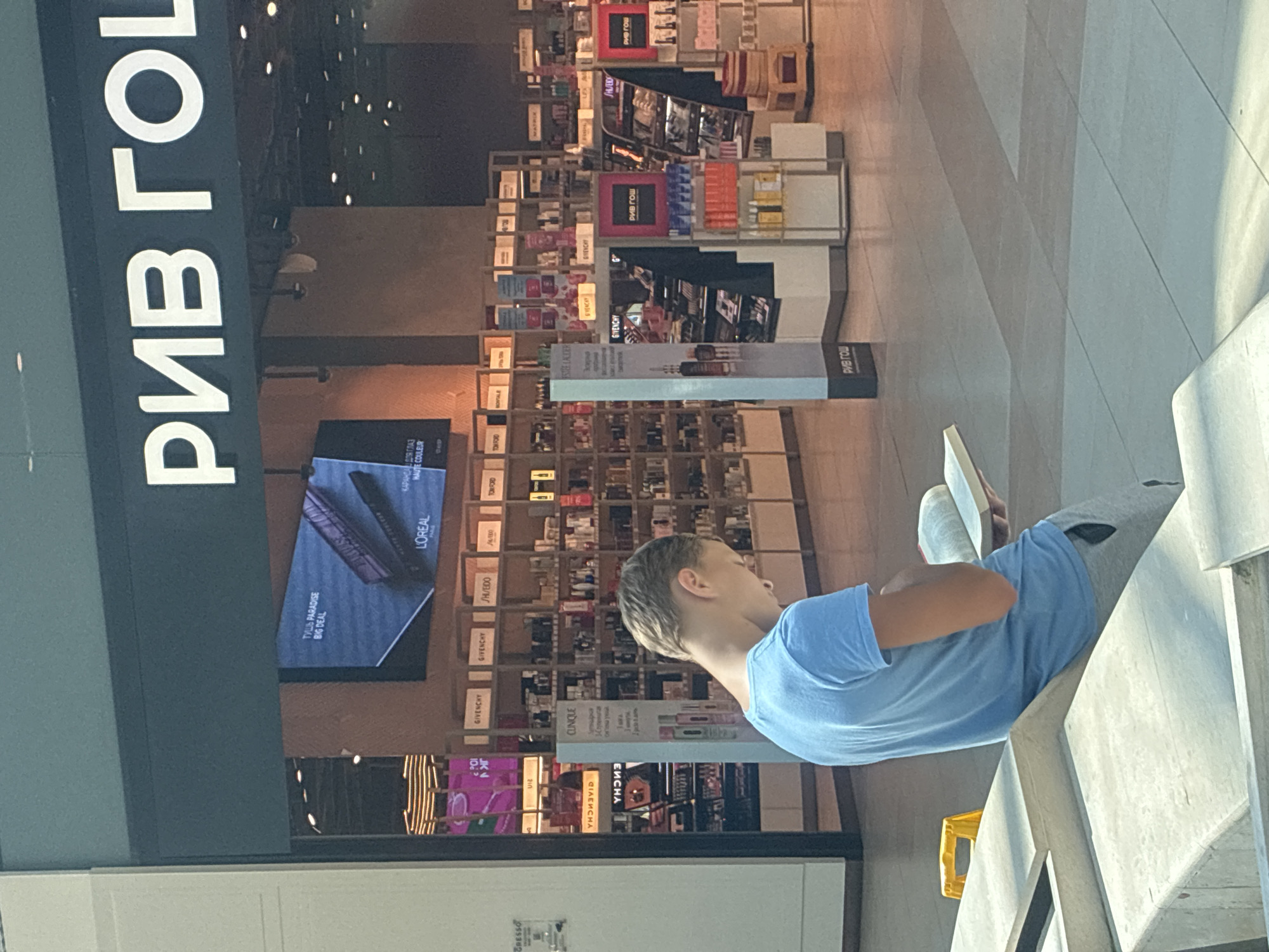 The image shows the interior of a large cosmetics store, likely a RIV GOSH store, located in Sochi, Russia. In the foreground, a young boy with blond hair, wearing a light blue t-shirt and grey sweatpants, is seated and reading a book. He is facing away from the camera, to the left. Behind him, the store is filled with shelves displaying a variety of cosmetic products. Brands such as L'Oréal, GIVENCHY, SHISEIDO, and TOM FORD are visible on signage and product packaging. The store features bright lighting and a clean, modern aesthetic. A digital screen is mounted above the merchandise, displaying an advertisement for L'Oréal mascara. The floor is tiled in a neutral color, and the overall environment suggests a shopping mall or a large retail space.