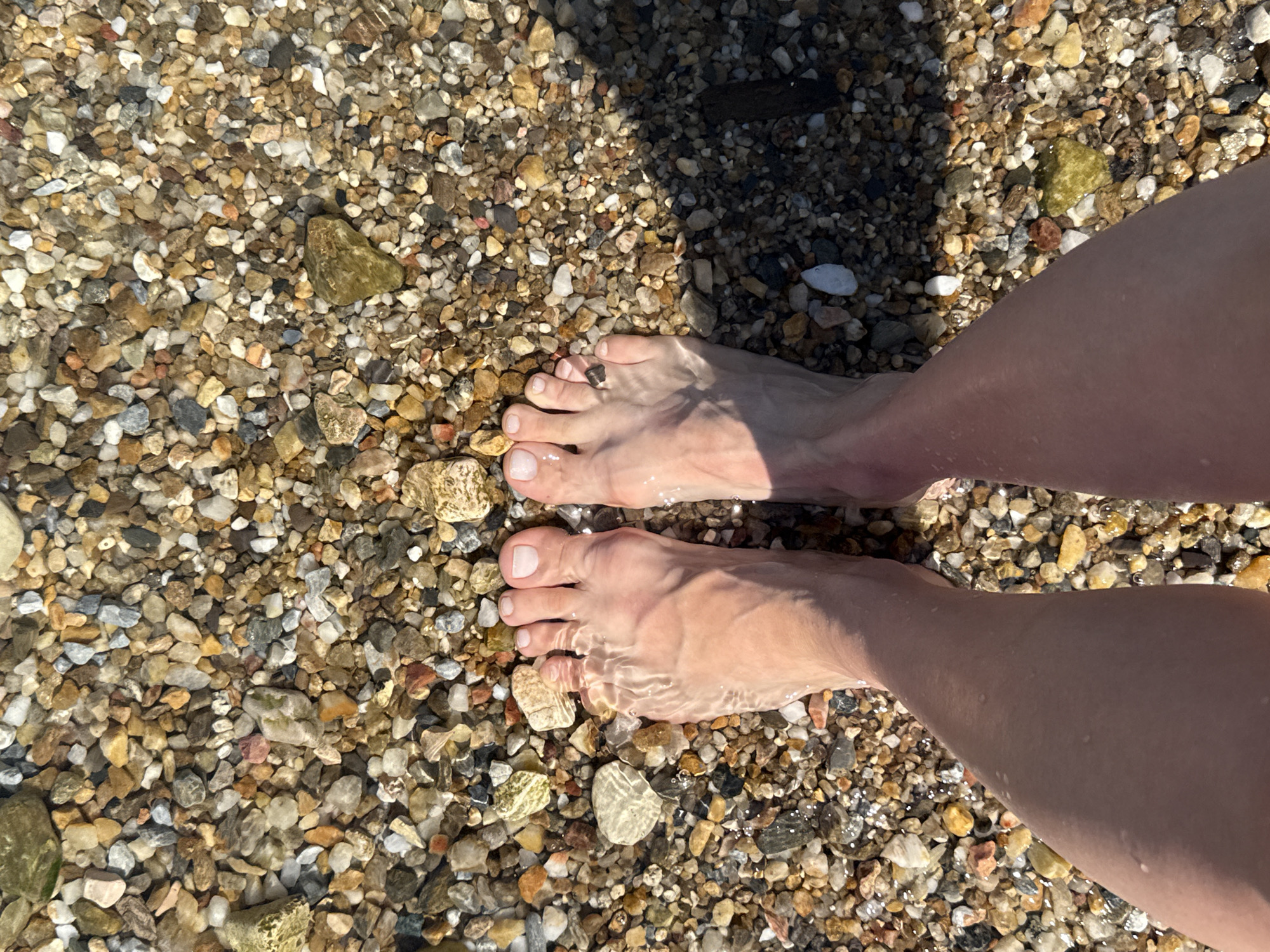 In Bodrum, Turkey, a person's bare feet are submerged in shallow, clear water on a pebbled beach. The pebbles are varied in size, color, and texture, ranging from small grains to larger stones in shades of white, grey, brown, yellow, and green. Sunlight creates dappled patterns and reflections on the water and the skin of the feet. The image is a top-down perspective, focusing on the feet and the immediate surroundings of the beach.