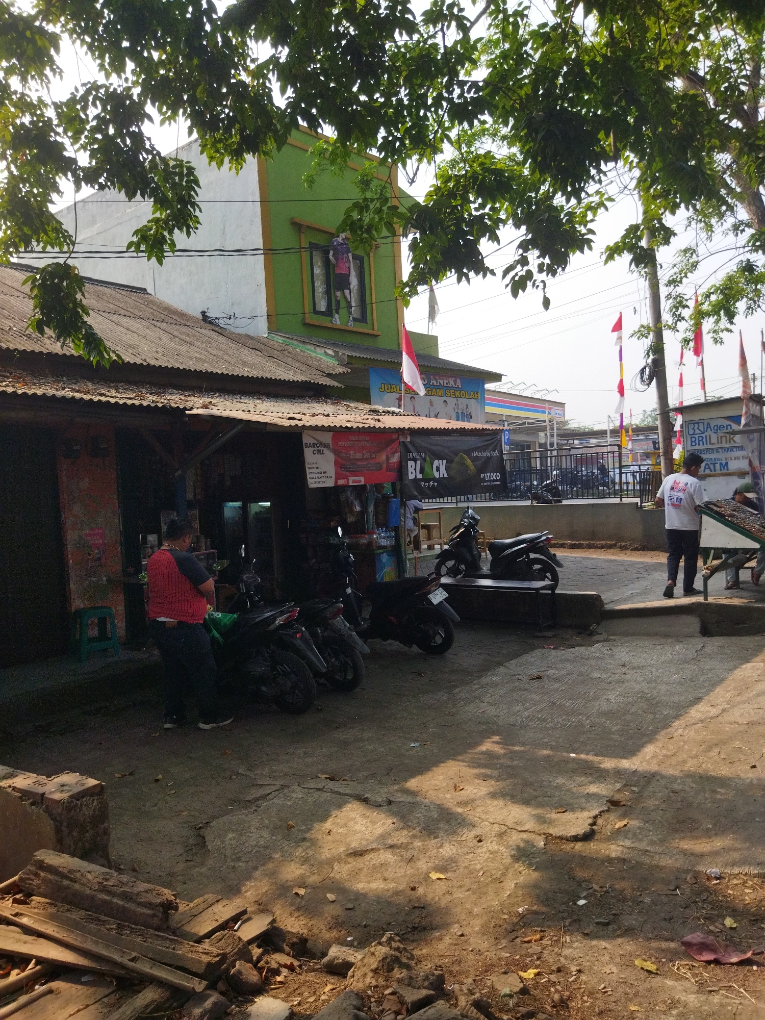 The image depicts a street scene in Sepatan, Indonesia. In the foreground, a man in a red and black shirt stands near a green stool in front of a building with a corrugated metal roof. Several motorcycles are parked in front of the building. To the right, another man in a white shirt walks away from the camera towards a table with items on display. In the background, a green and white building with a mural of a person in athletic wear is visible. Various flags are strung across the scene. The ground is paved with concrete and dirt, with scattered debris. The overall impression is that of a commercial area with an informal marketplace.