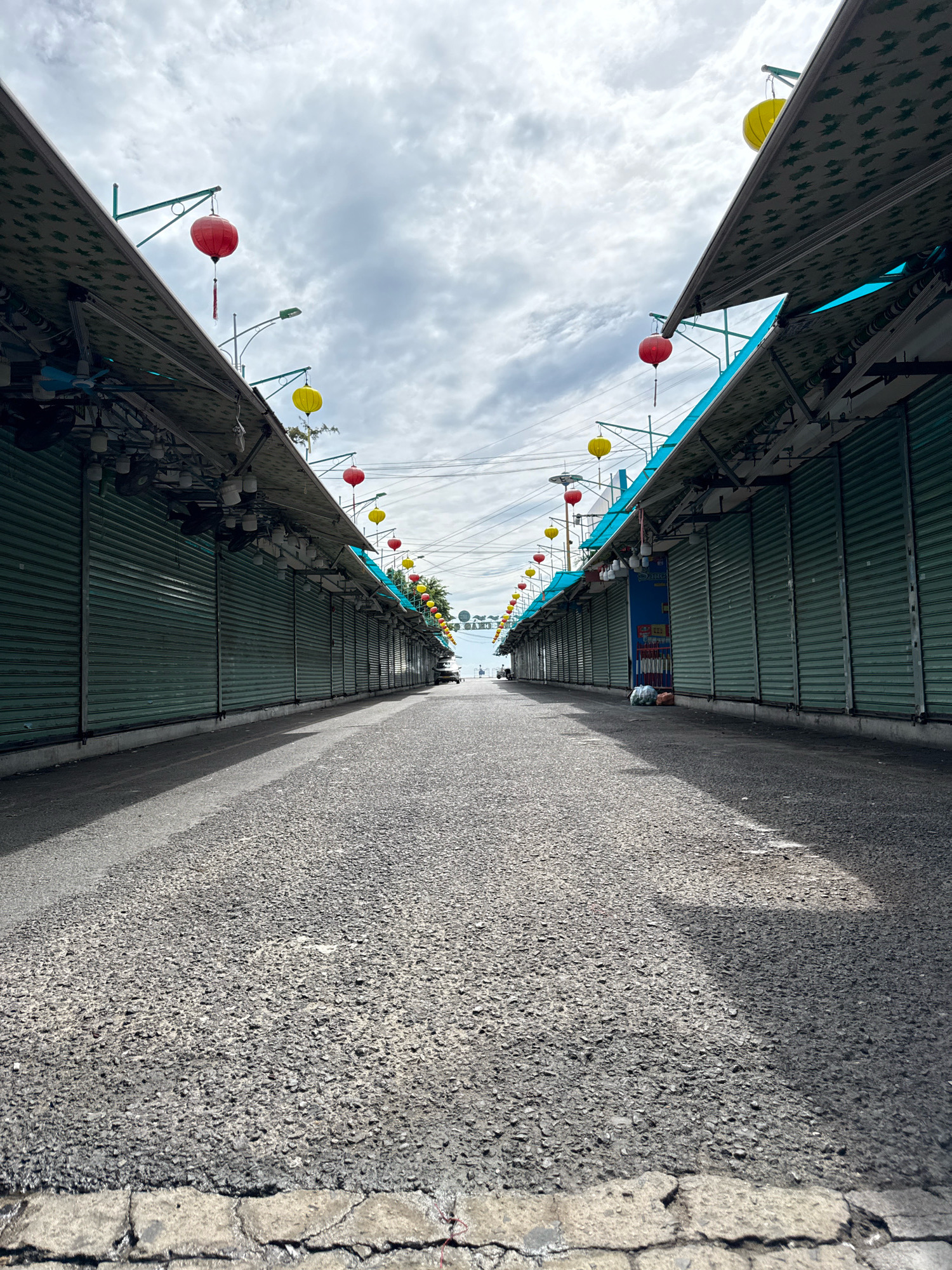The image depicts an empty street lined with closed market stalls in Nha Trang, Vietnam. The stalls have green metal roll-up shutters, and their awnings are adorned with green fabric featuring a white patterned design. Red and yellow lanterns are suspended from poles above the stalls, adding a touch of color to the otherwise subdued scene. The street is paved with asphalt and shows signs of wear. The sky is overcast with clouds, suggesting a potentially humid or rainy day. The perspective is low, looking down the street from ground level, emphasizing the symmetry of the stalls and the long, empty road stretching into the distance. A few distant objects, possibly vehicles or people, are visible at the far end of the street, near where the road meets the sky. The overall impression is one of quietness and inactivity, perhaps indicating an early morning, a closure period, or a time of low foot traffic.