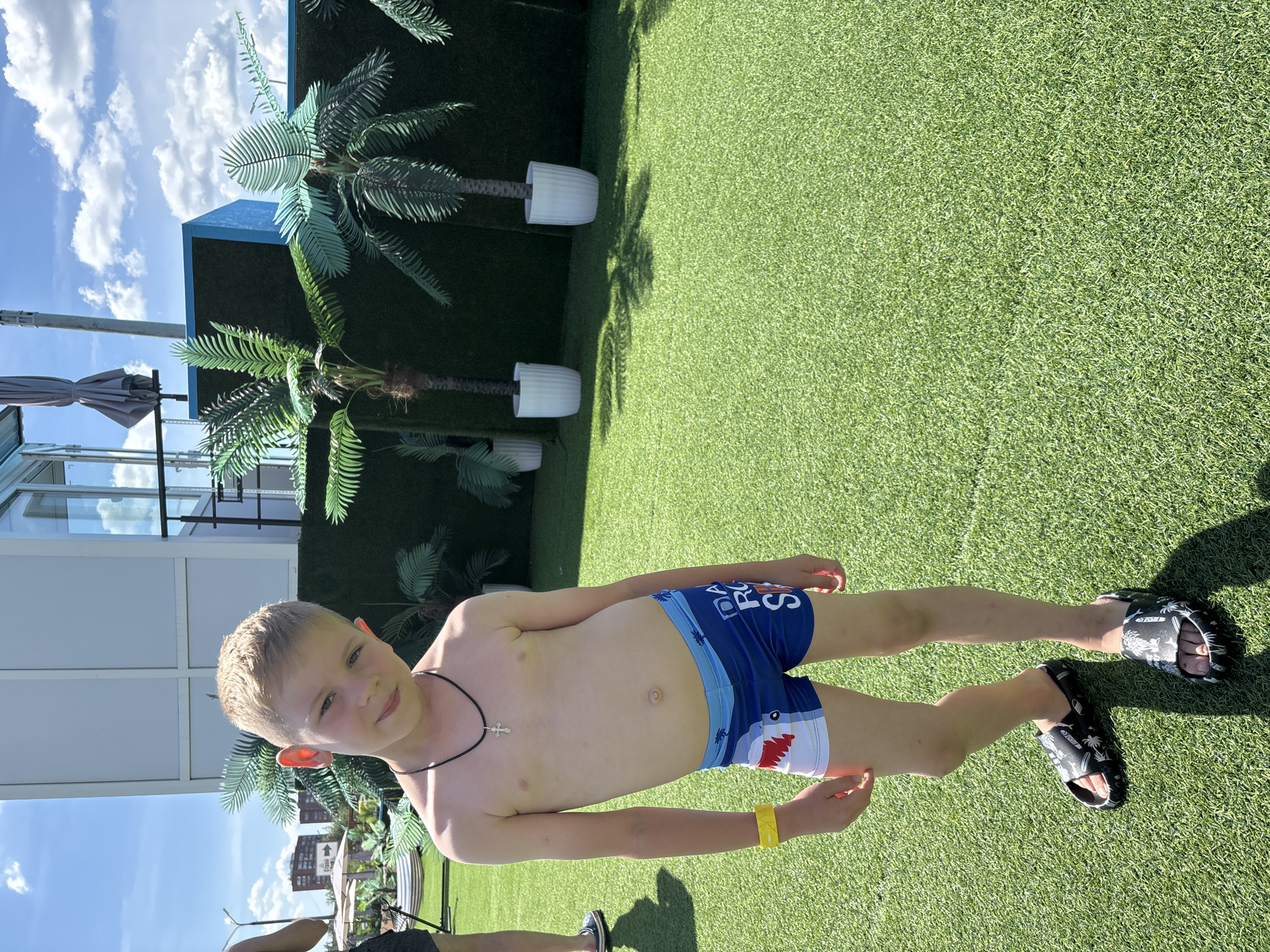 A young boy with blonde hair and light skin is standing on a green, artificial turf surface. He is wearing blue swimming trunks with a shark design and the words "DIAL FOR S..." visible, a black necklace with a cross pendant, and a yellow wristband. He has dark shoes with a white pattern. He is looking towards the camera with a slight smile. The background features artificial palm trees in white pots, a blue structure, and a building with large windows. The sky is blue with white clouds, suggesting it is a sunny day. The overall scene appears to be an outdoor recreational area, possibly a water park or resort. The image is taken in Yaroslavl, Russia, during daytime.