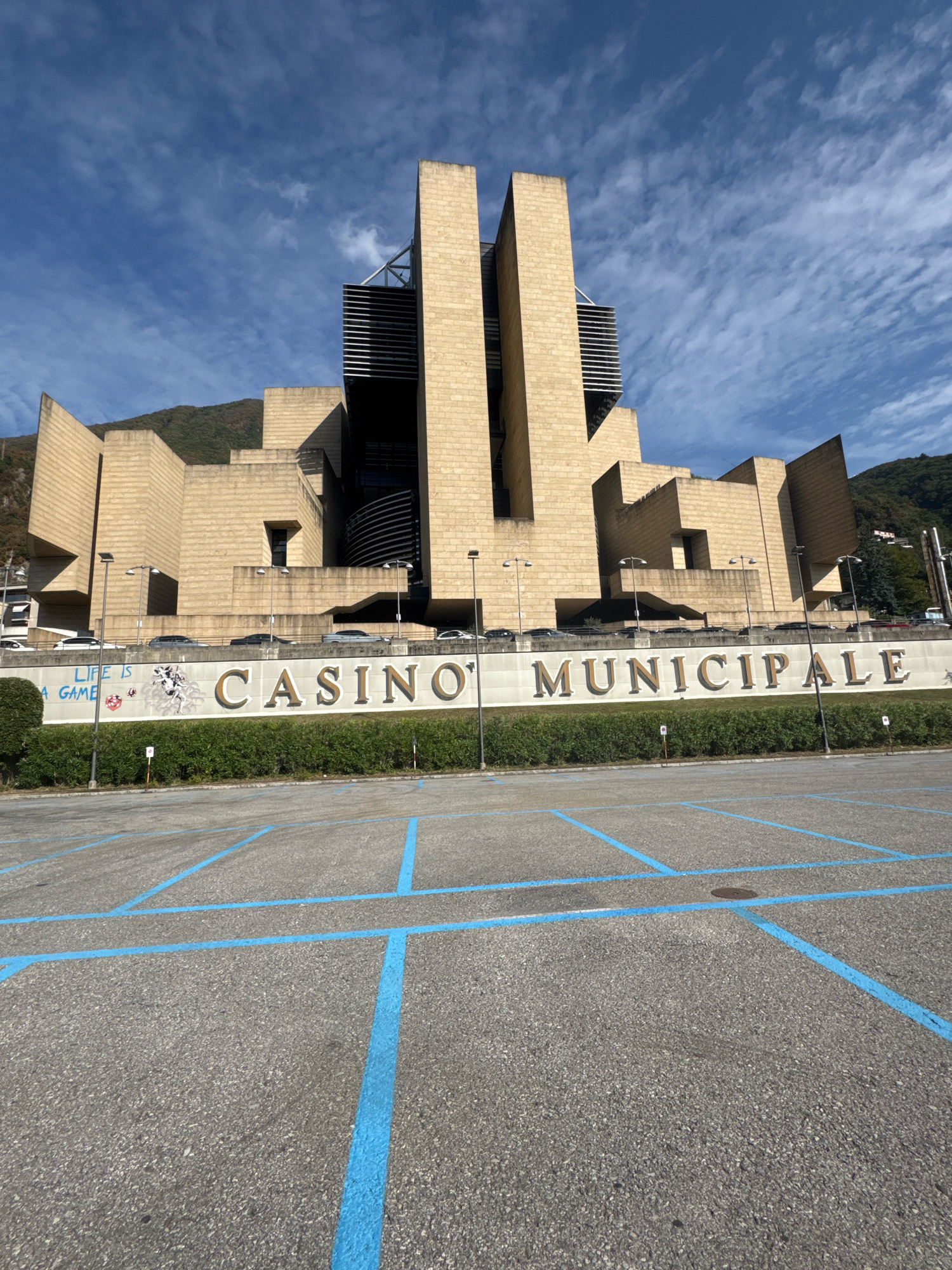 The image shows the Casino Municipale in Campione d'Italia, Italy. The building has a distinctive modern architectural style with angular, stepped sections and a light-colored stone facade. In the foreground, there is a paved parking lot marked with blue lines. Behind the parking lot, a low wall with a hedge runs along the front of the casino. Large gold lettering on the wall spells out "CASINO MUNICIPALE". Below the word "CASINO", there are some graffiti tags, including the text "LIFE IS A GAME" and an image of a stylized animal head. Several cars are parked on a road or elevated platform behind the wall. Mountains covered in vegetation form the backdrop to the building under a blue sky with scattered white clouds.