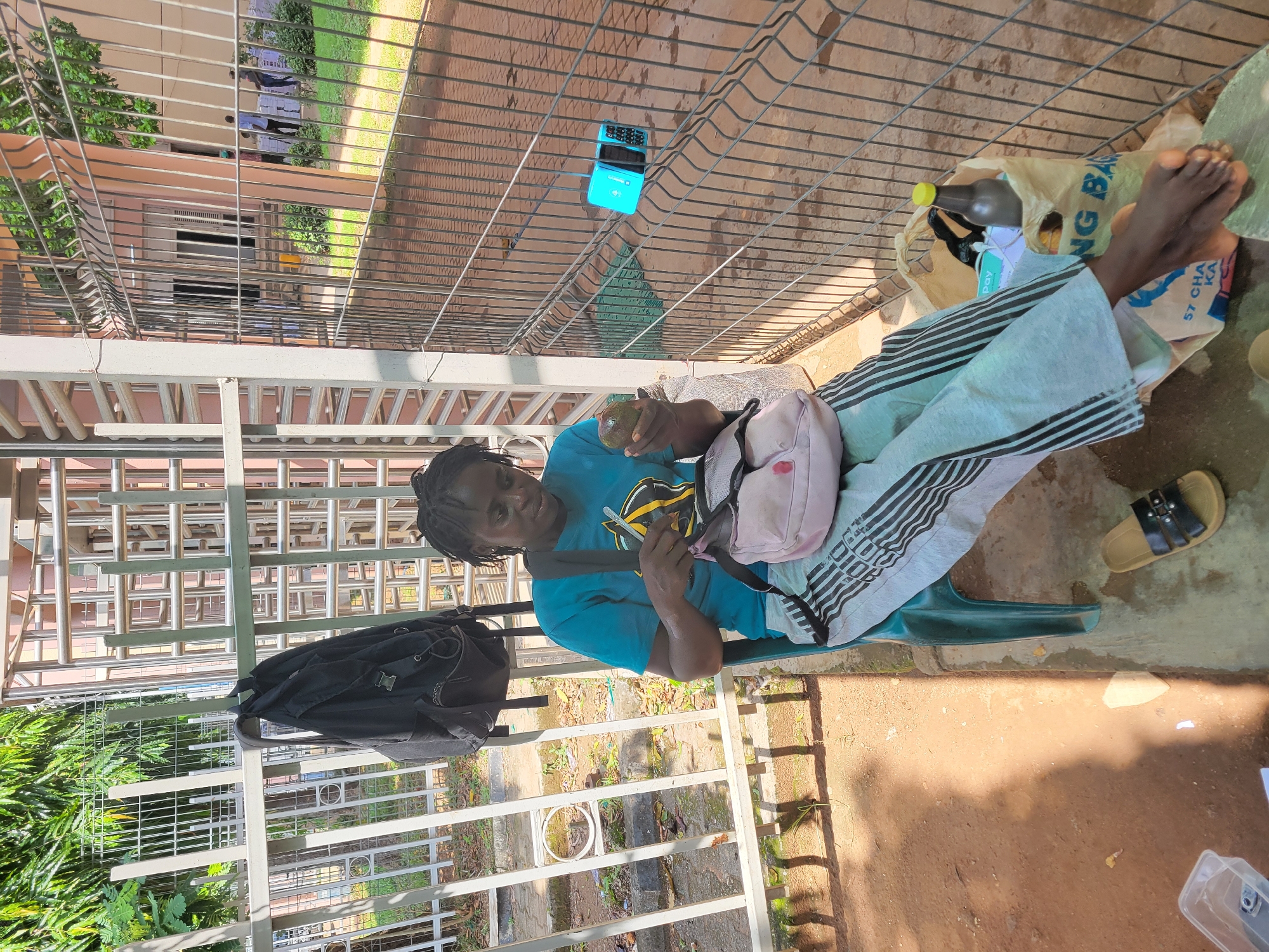 A woman is seated outdoors on a green plastic chair, likely in a shaded or semi-shaded area next to a building. She has dark skin and her hair is styled in dark braids or dreadlocks. She is wearing a short-sleeved turquoise t-shirt with a yellow and black graphic, and light-colored trousers with black horizontal stripes.
In her left hand, she holds a green, spherical fruit resembling an avocado. Her right hand appears to be adjusting something within a small pink shoulder bag that rests on her lap. To her left (viewer's right), a black backpack hangs from a metal structure or fence. Her right foot is bare, and a pair of tan and black sandals are placed on the reddish-brown ground nearby. A clear plastic bag containing a bottle with a yellow cap and other items is visible next to her feet. Further on the ground, a blue flip phone or small electronic device is present.
The setting appears to be an outdoor space, possibly a patio or walkway. A prominent metal fence, featuring both vertical and horizontal bars, stretches across the foreground and midground, separating the immediate area from a lush green space with trees and shrubs. Beyond this, light-colored buildings with windows are visible under bright daylight, casting distinct shadows. The ground is composed of reddish-brown earth or concrete with scattered debris.
The time of day is bright, likely midday or afternoon, given the strong illumination. The woman's expression is neutral, suggesting she is calmly engaged in her activity. Visible text on the plastic bag includes "NG BA," and the word "Pay" can be seen on an item within the bag. The scene conveys a relaxed atmosphere in what appears to be a warm climate.