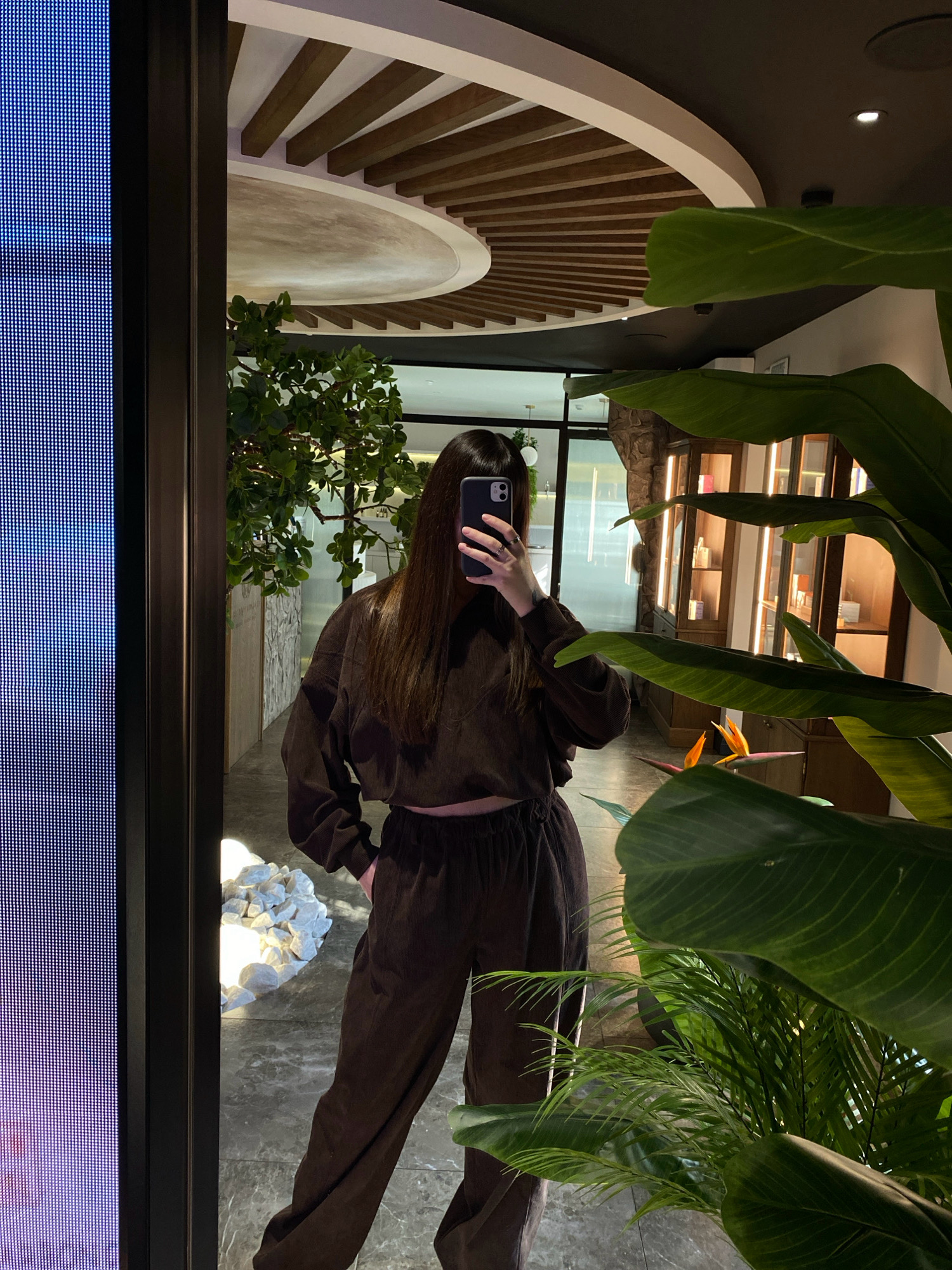 A young woman with long, dark hair is taking a mirror selfie in what appears to be a boutique or retail space. She is wearing a brown corduroy two-piece set, consisting of a long-sleeved top and loose-fitting pants. She is holding a smartphone in her right hand, obscuring her face.
The background reveals a modern interior with a large, circular ceiling feature with wooden slats. To the left of the woman, a large digital display screen with blue pixels is visible. To the right, lush green plants, including large-leafed tropical varieties and a palm plant, are strategically placed, adding to the ambiance. In the middle ground, there's a display of decorative white stones. Further back, glass-fronted cabinets with illuminated shelves suggest a retail display, possibly for cosmetics or perfumes, and a stone-clad wall element is visible. The floor is tiled with a marble-like pattern.
The location is identified as Votkinsk, Russia.