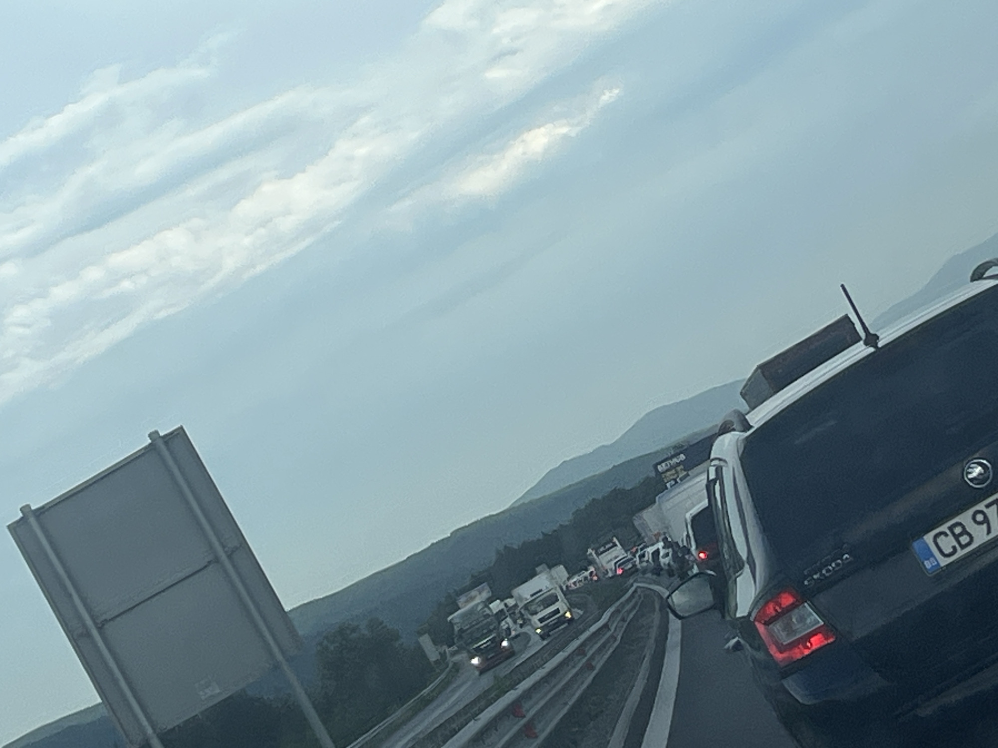 The image depicts a traffic jam on a highway near Vakarel, Bulgaria. The sky is overcast with scattered clouds. In the foreground, a black Skoda car is visible, with its license plate reading "CB 97 XX". Further ahead, several trucks and cars are stopped on the highway. The road appears to be a multi-lane highway with concrete barriers. In the background, rolling hills covered in trees are visible, suggesting a rural or semi-rural setting. The overall impression is one of congestion and slow-moving traffic.