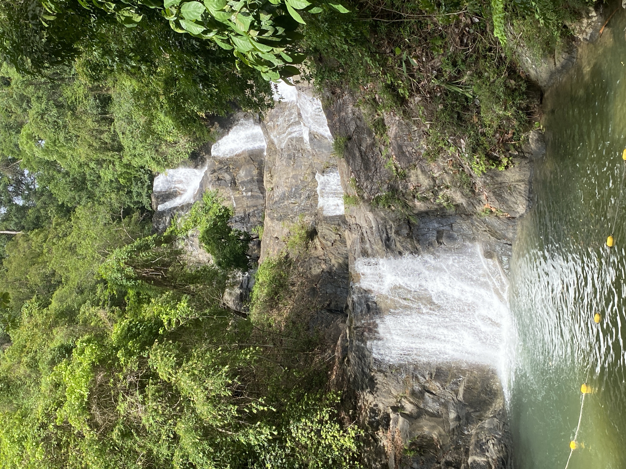 The image depicts a tiered waterfall cascading down a rocky cliff face into a pool of water, surrounded by lush green vegetation. The location is identified as Tambon Thai Mueang, Thailand. The water flows down multiple levels, creating a series of small falls. The pool at the bottom is lined with a rope and yellow buoys. The surrounding area is densely covered with trees and plants, growing on both sides of the waterfall.