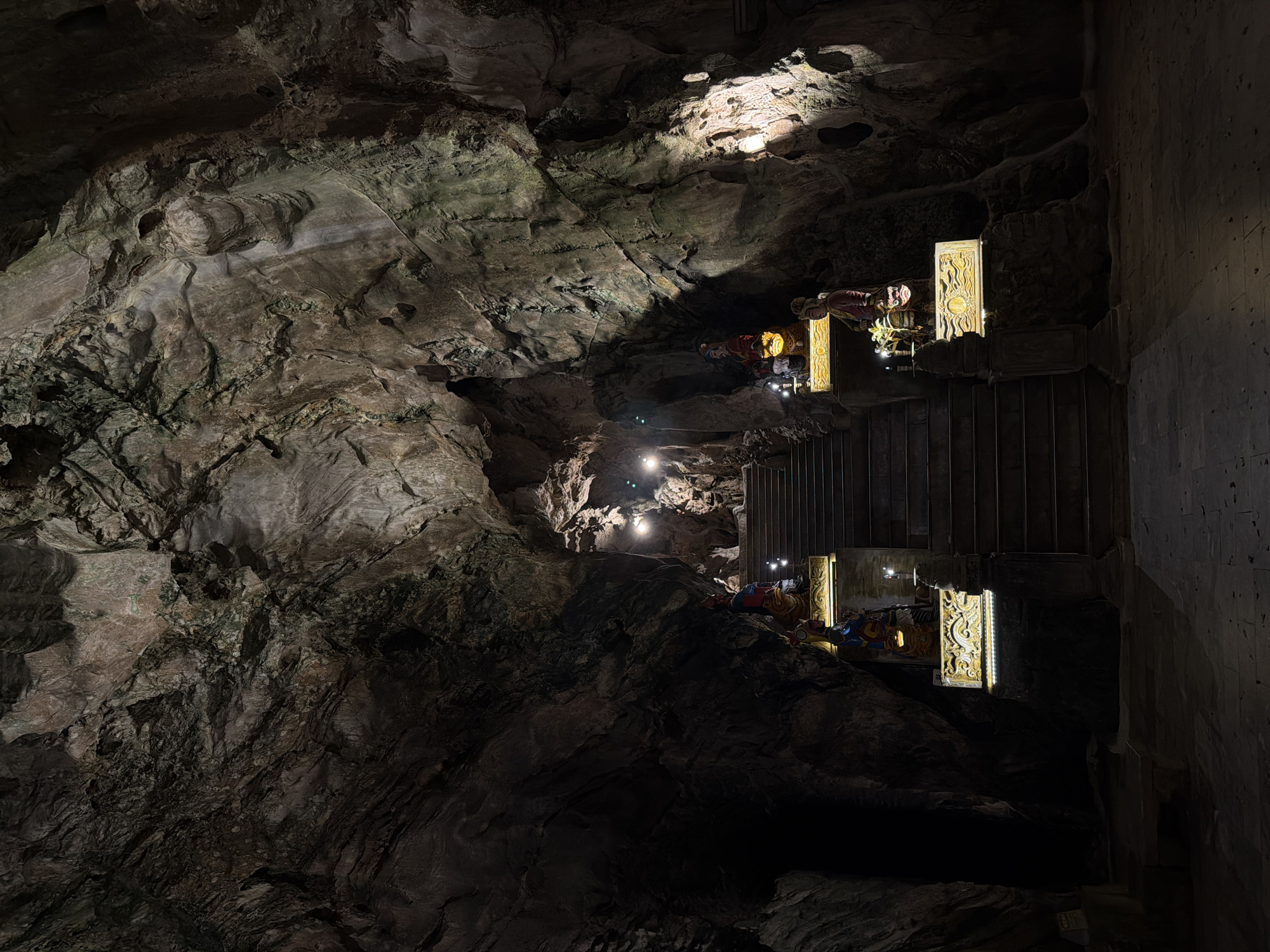 The image shows the inside of the Marble Mountains cave system near Đà Nẵng, Vietnam. The cave walls are rough and uneven, with textured rock formations. Several people wearing helmets and colorful clothing are visible along a pathway with artificial lighting. Ornate decorative elements, possibly part of a temple or shrine, are positioned along a set of stairs. A portion of the cave wall is visible on the right side.