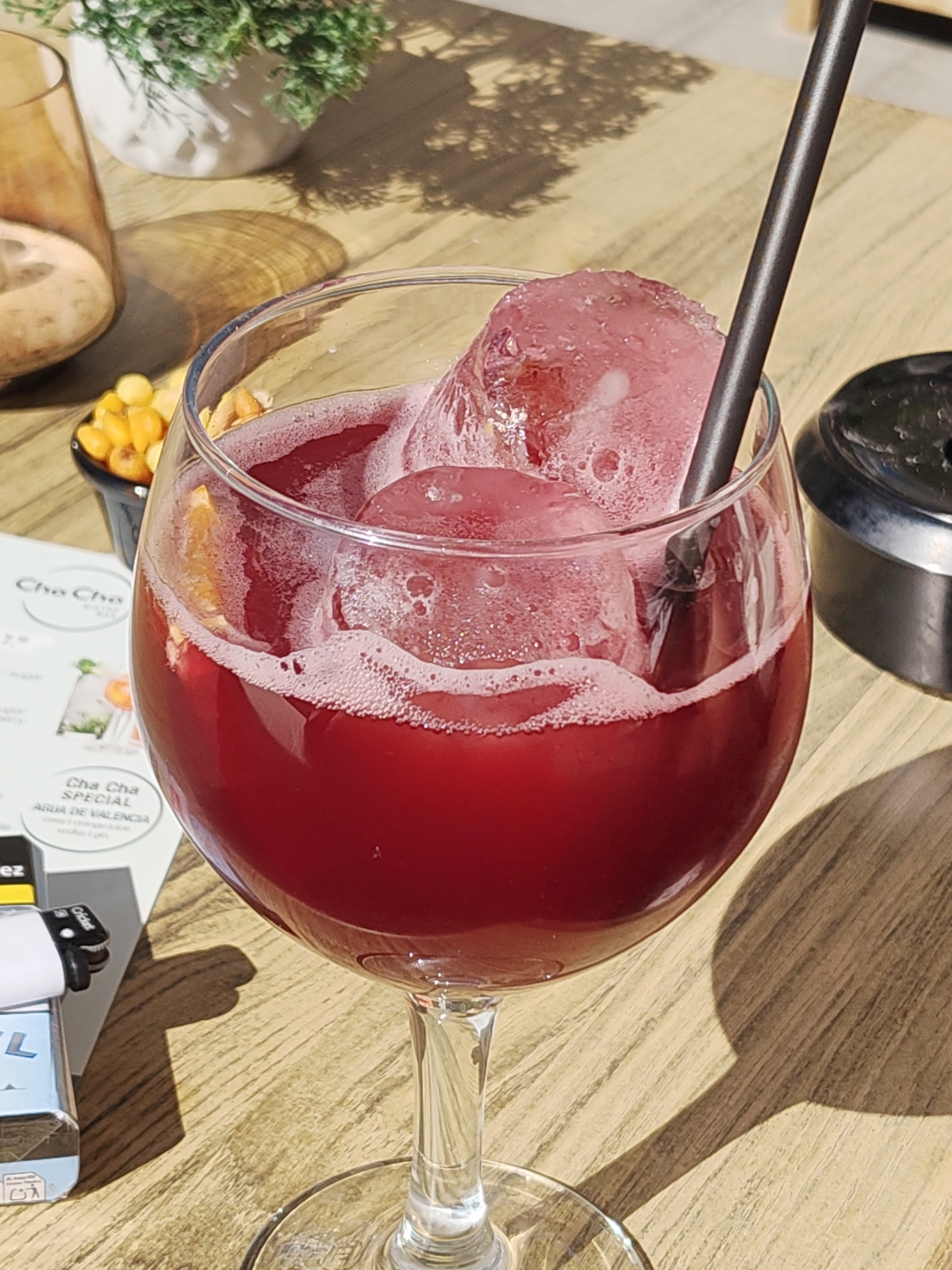 A close-up captures a large glass of red wine, brimming with ice, possibly sangria, and a dark straw on a wooden tabletop. The foam from the drink is visible, along with small orange pieces in the wine. Beside the glass, there is a small bowl of yellow berries and a metallic ashtray. A menu or promotional material with the words "Cha Cha Special Agua de Valencia" is partially visible. A lighter and cigarette pack are located to the bottom left. The scene is outdoors, likely in the city of Dénia, Spain, indicated by the text on the menu. The lighting suggests it's daytime. No people are visible.