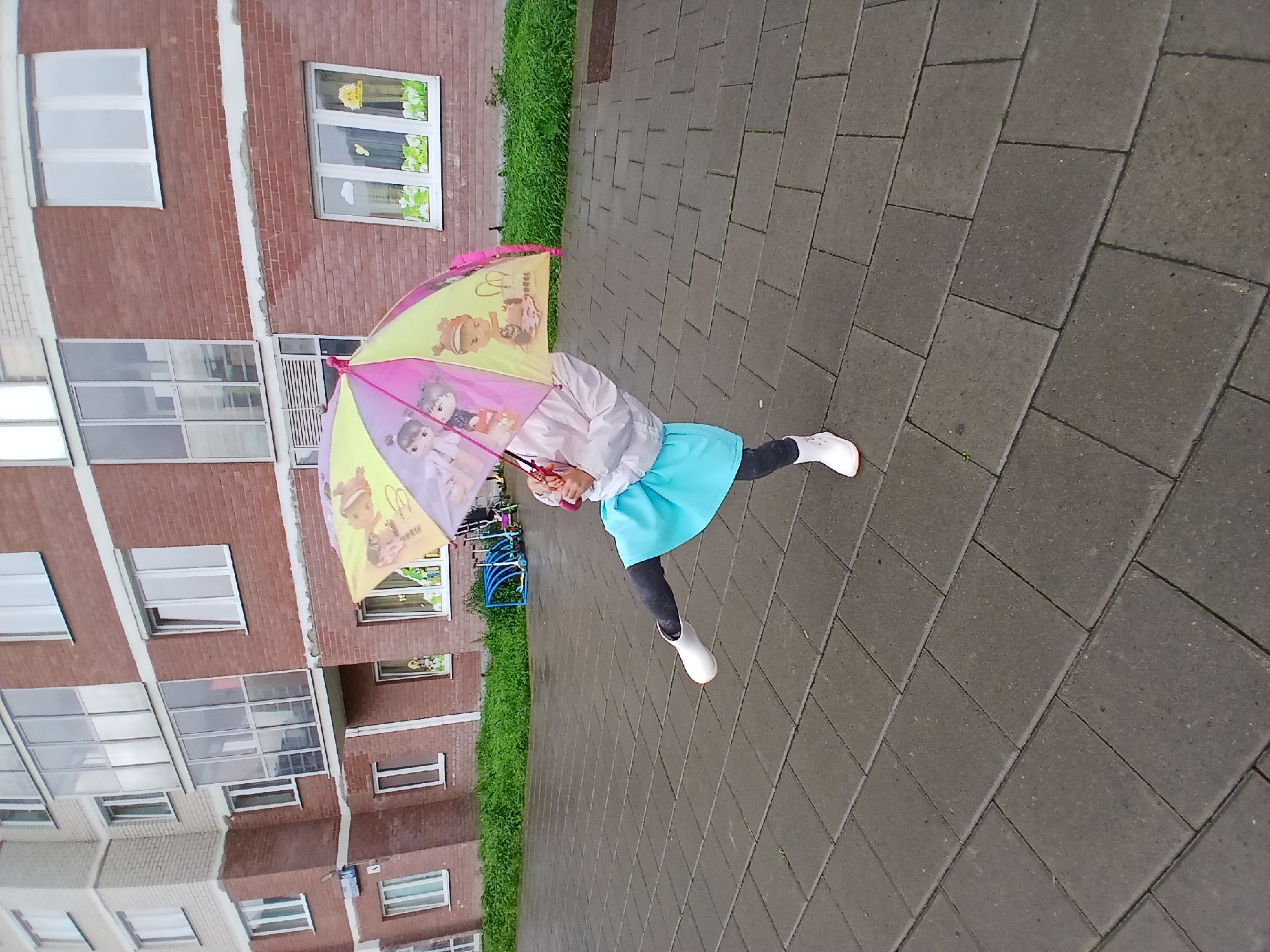 A young girl is standing on a paved area in front of a brick apartment building, holding an open, multi-colored umbrella featuring cartoon characters. She is wearing a light-colored jacket, dark leggings, and white boots. The umbrella is tilted, and her body is angled as if she is in motion. The building has multiple windows, and a balcony or enclosed porch is visible. Green bushes line the base of the building. In the background, blue scooters are visible. The ground is paved with rectangular grey stones. The scene appears to be in an urban environment, possibly a residential courtyard in Yekaterinburg, Russia, on a cloudy day.